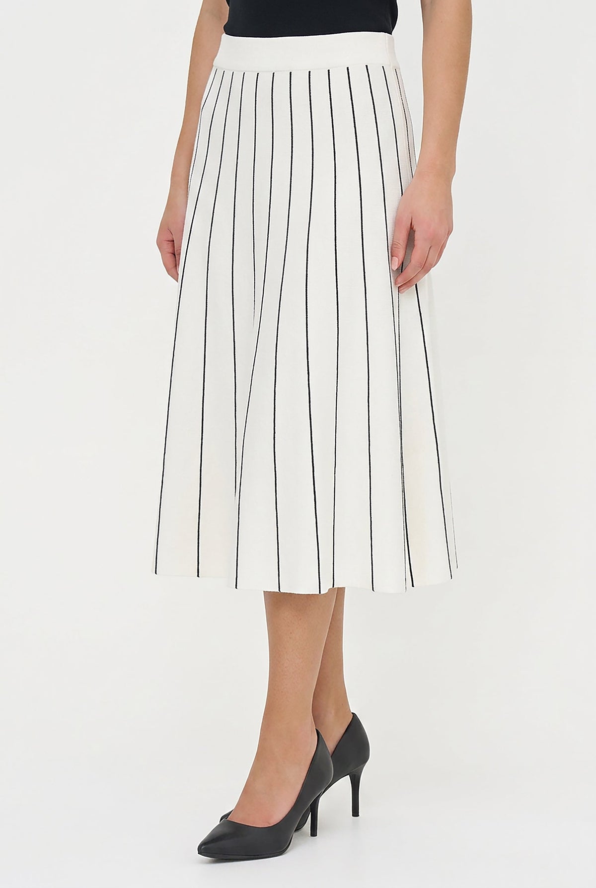 A model wearing a white pleated midi skirt with thin black vertical stripes, paired with a black top and black pointed-toe heels, against a plain white studio background.