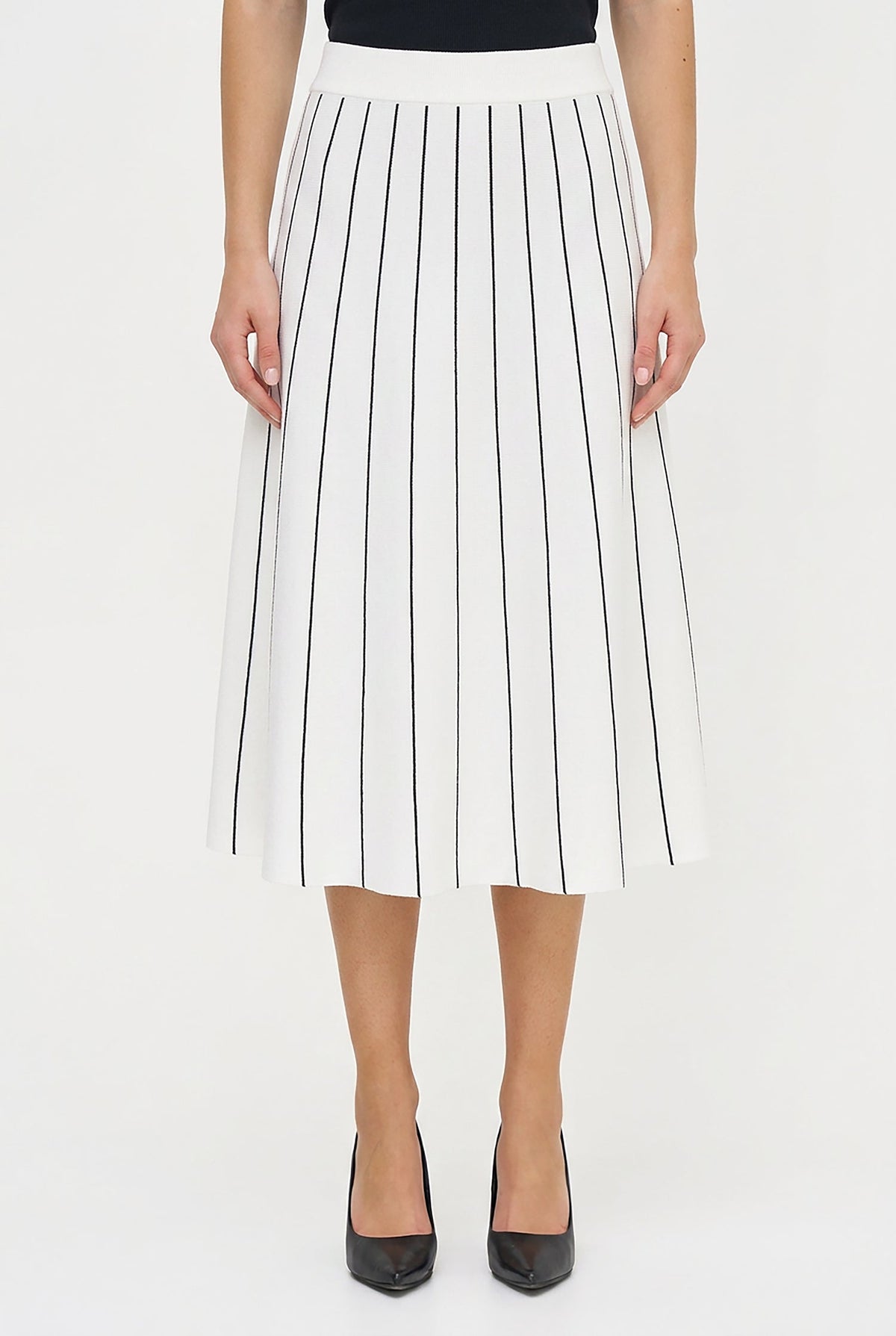 A model wearing a white pleated midi skirt with thin black vertical pinstripes, styled with a black top and black pointed-toe heels against a plain white studio background.