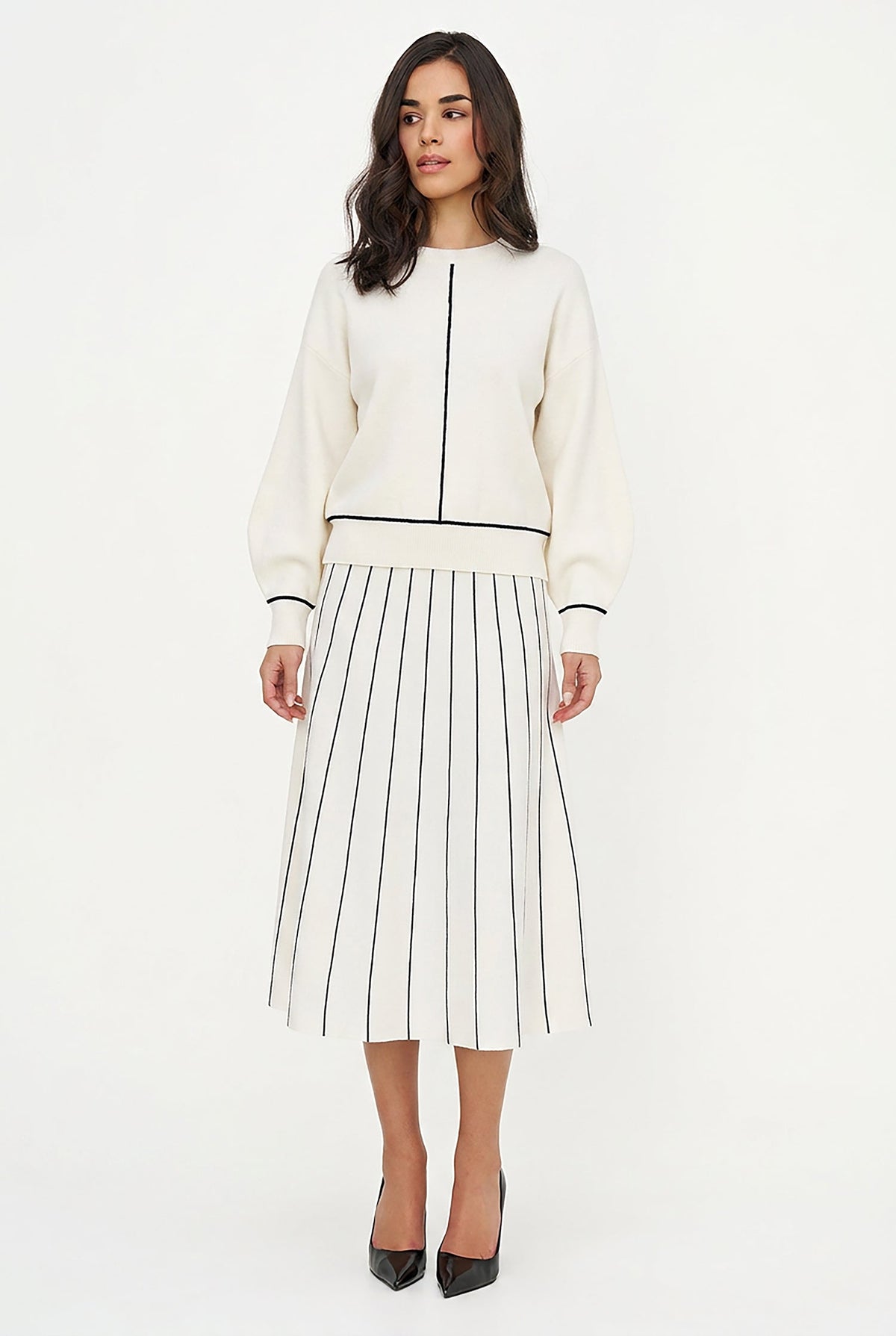 A woman modeling a cream-colored knit sweater and matching pleated midi skirt set with thin black stripes and black trim details, paired with black pointed-toe heels, against a plain white studio background.