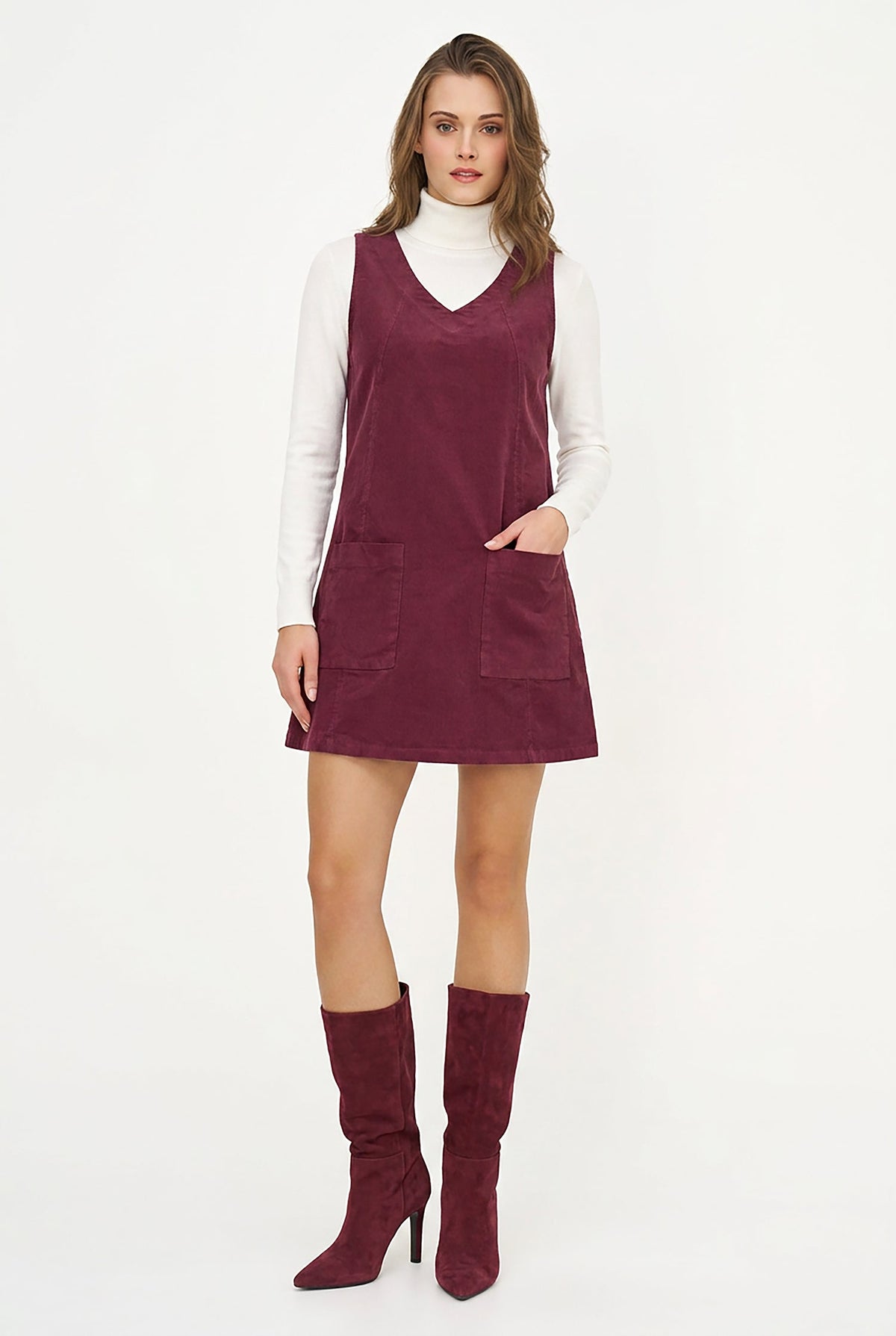 A woman modeling a sleeveless burgundy corduroy pinafore mini dress over a white turtleneck, paired with matching burgundy heeled boots, on a white studio background.