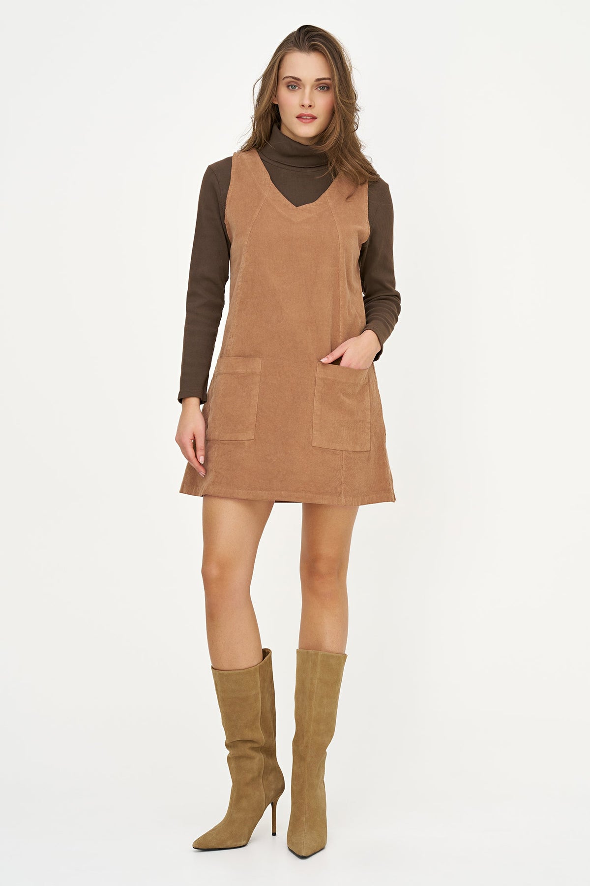 A woman modeling a brown corduroy pinafore dress over a dark brown turtleneck, paired with tan knee-high heeled boots, against a plain white studio background.