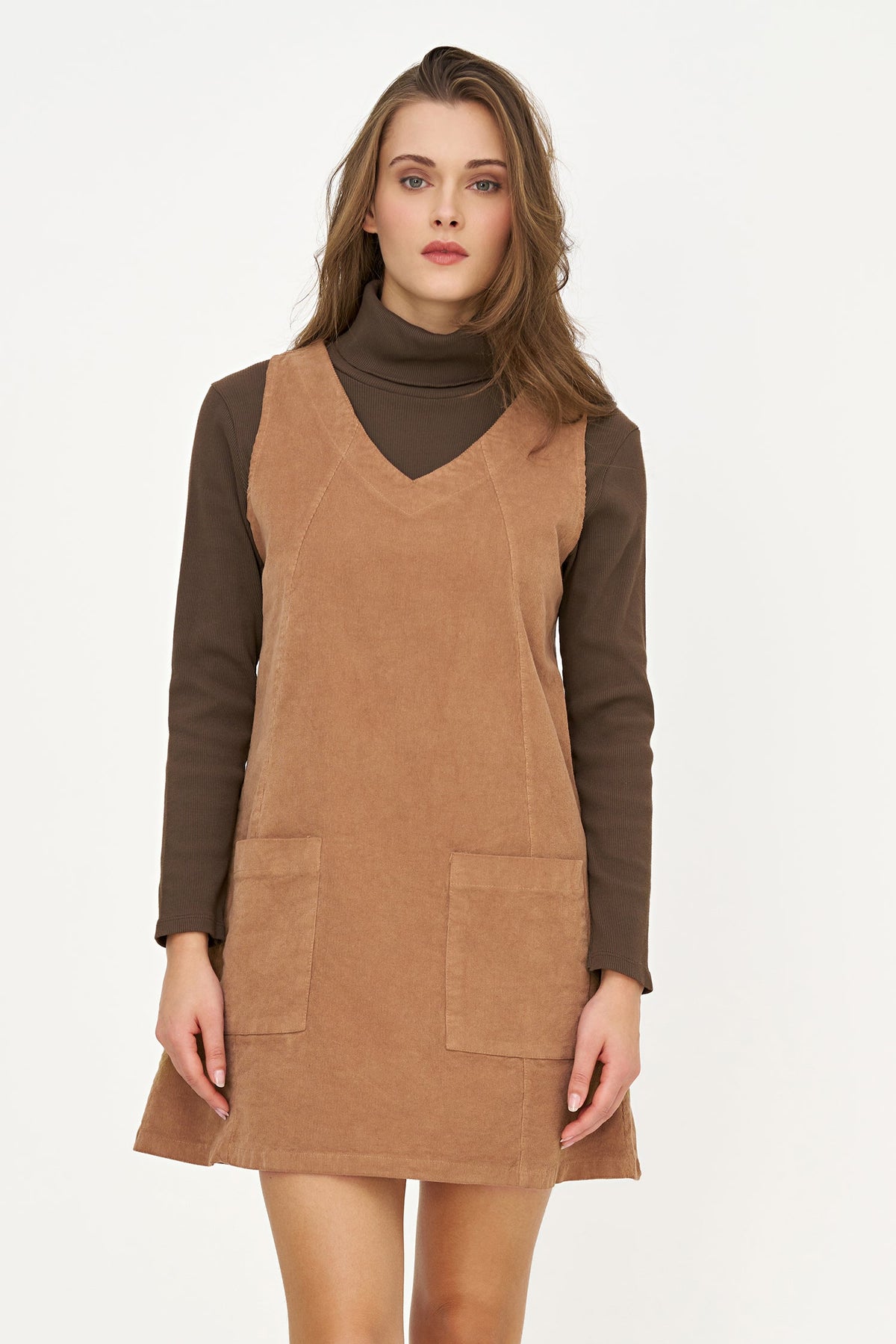 A woman modeling a brown corduroy pinafore dress over a dark brown turtleneck sweater, photographed against a plain white studio background.