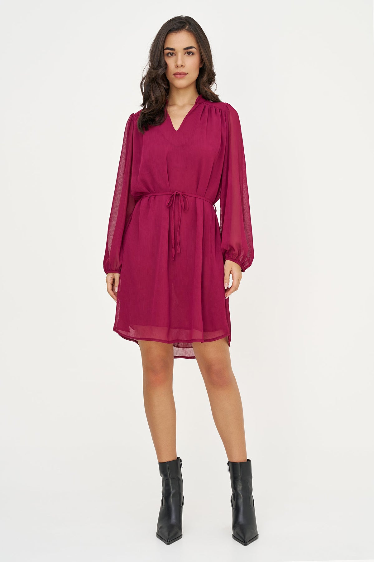 A woman modeling a magenta long-sleeve chiffon mini dress with a tie waist, paired with black ankle boots, against a plain white studio background.