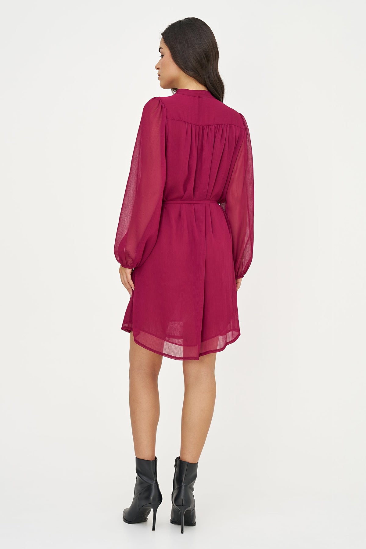 A woman viewed from the back wearing a sheer magenta long-sleeve mini dress with a gathered waist, paired with black ankle boots, against a plain white studio background.