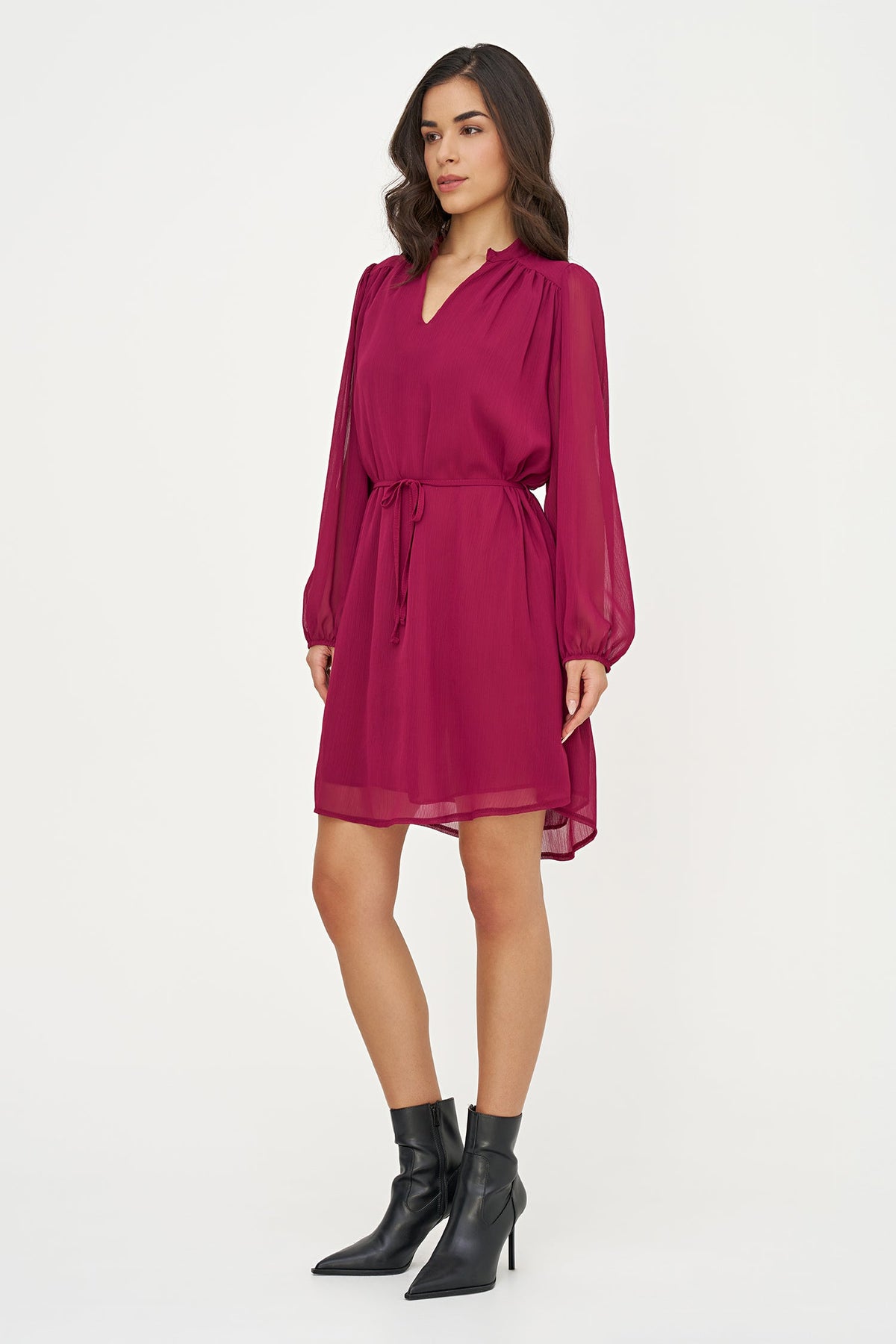 A woman modeling a long-sleeved magenta chiffon mini dress with a tie waist, paired with black pointed-toe ankle boots, against a plain white studio background.