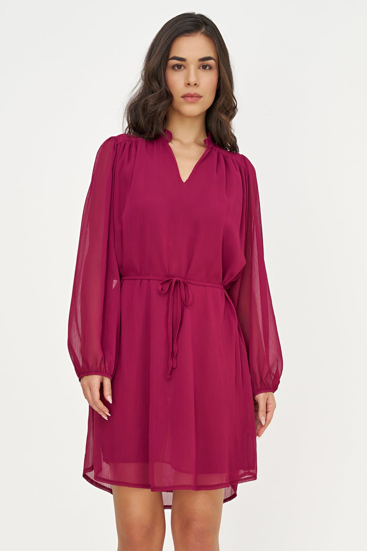 A woman modeling a magenta chiffon mini dress with long sheer sleeves, a V-neckline, and a matching fabric tie belt at the waist, set against a plain light gray studio background.