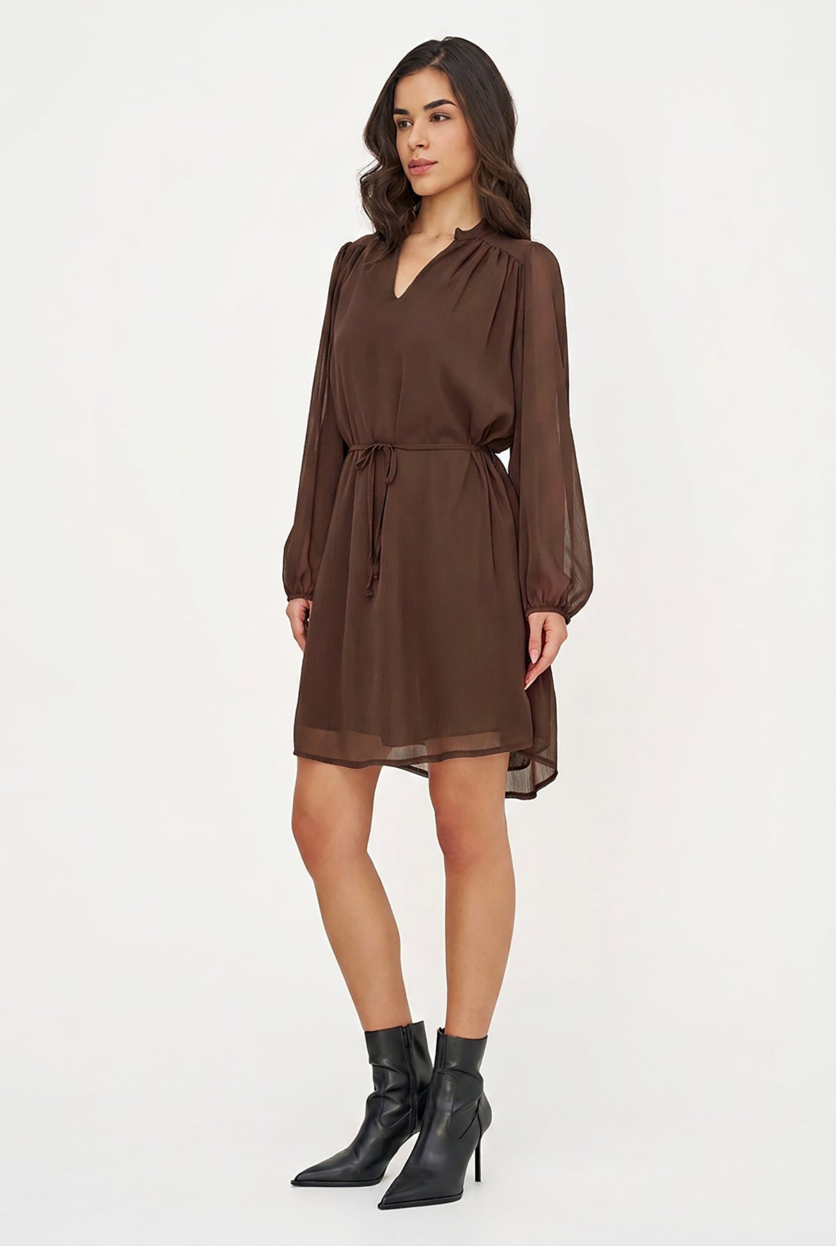 A woman modeling a brown long-sleeve mini dress with a V-neckline, gathered waist, and sheer balloon sleeves, paired with black pointed-toe ankle boots against a plain white studio background.