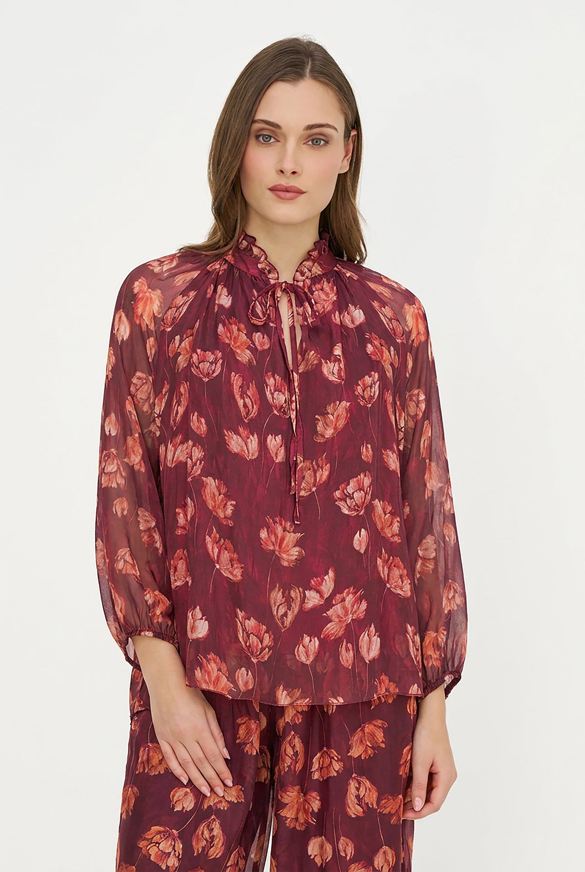 A woman modeling a sheer long-sleeve blouse and matching pants set in a deep burgundy color with a red and orange floral pattern, set against a plain white studio background.