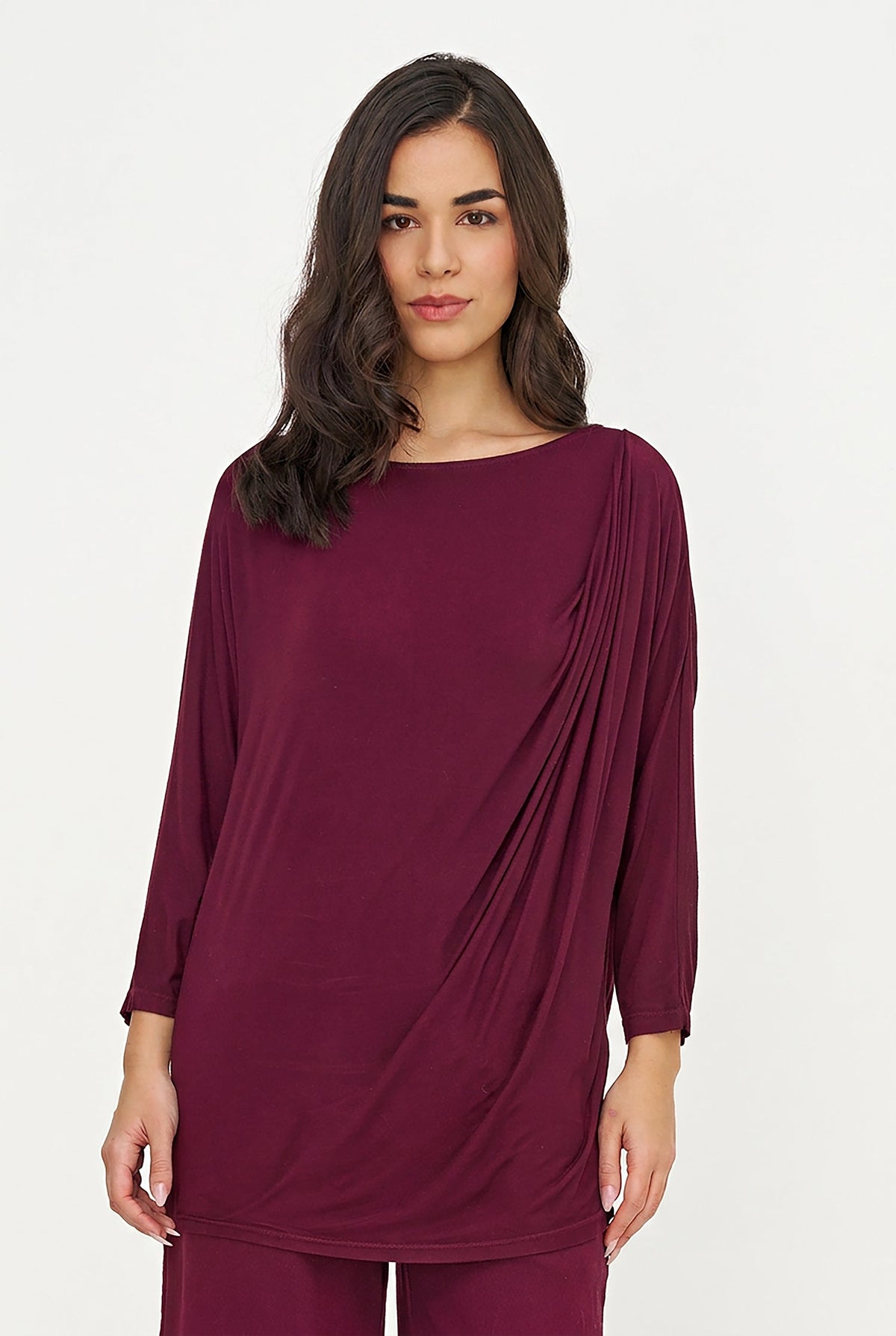 A woman modeling a draped long-sleeve tunic top in deep burgundy, paired with matching pants, against a plain white studio background.