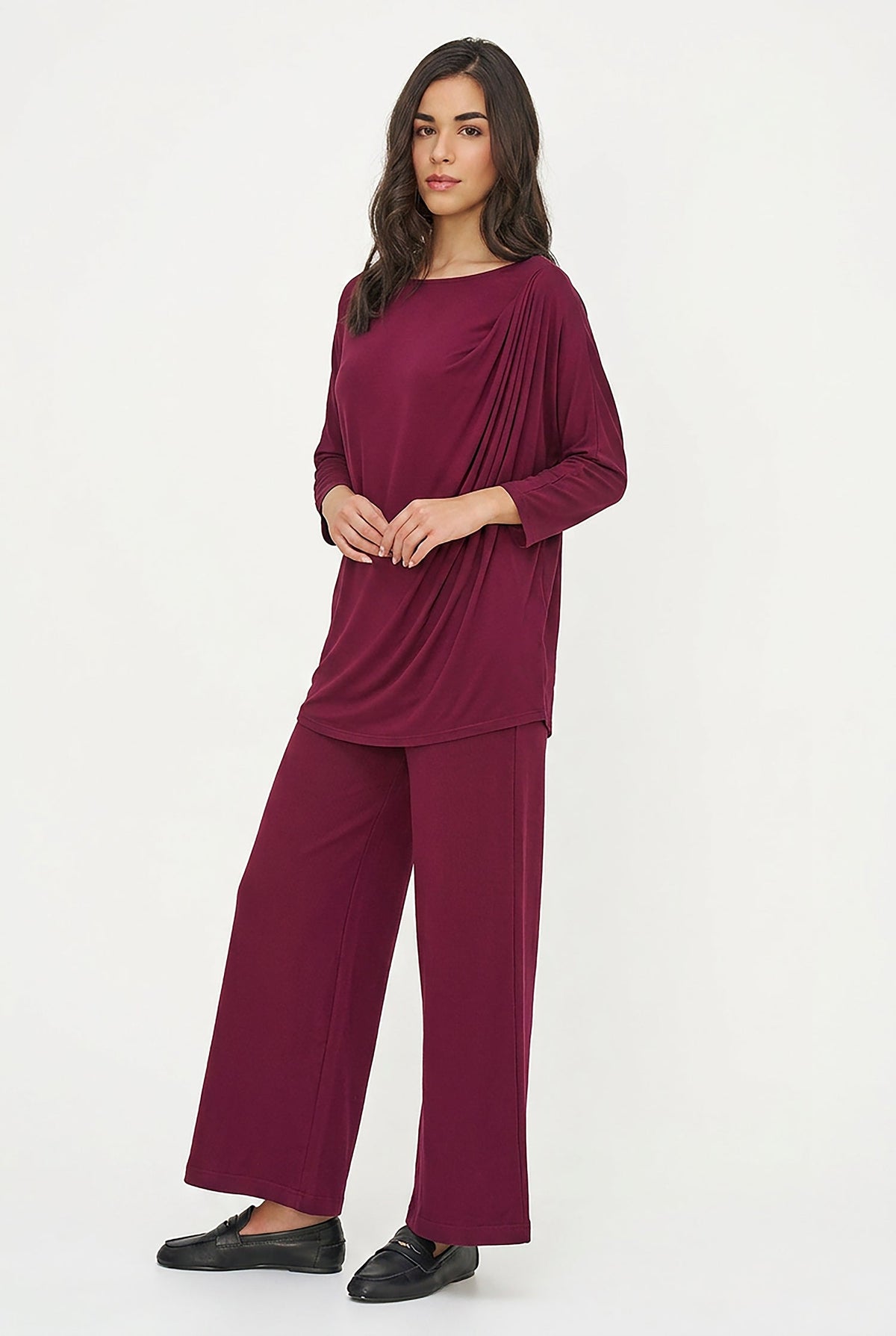A woman modeling a matching two-piece loungewear set in deep burgundy, featuring a draped long-sleeve top and wide-leg pants, paired with black loafers against a plain white studio background.
