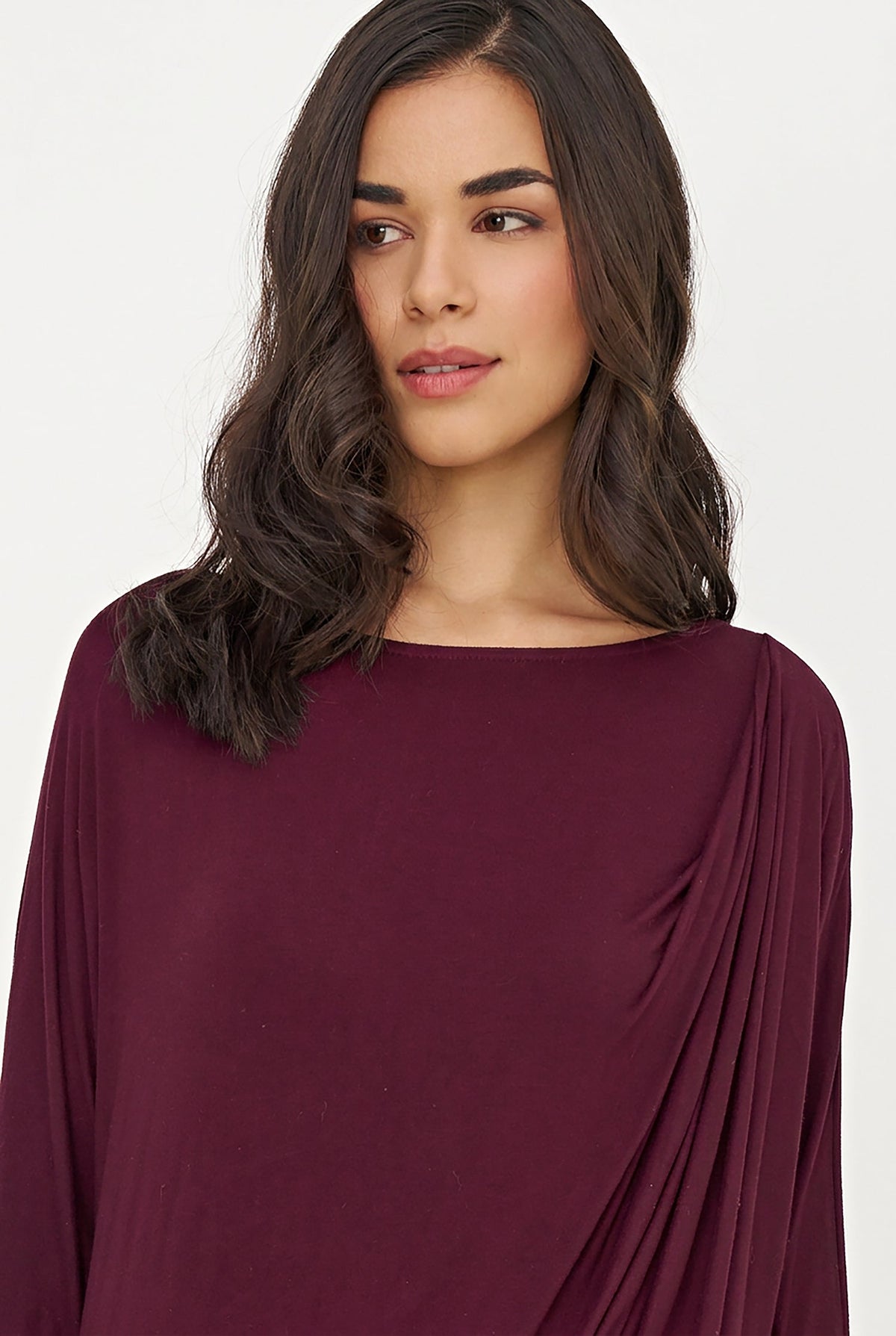 A woman with dark wavy hair wearing a draped, long-sleeve top in deep burgundy, photographed against a clean off-white background.
