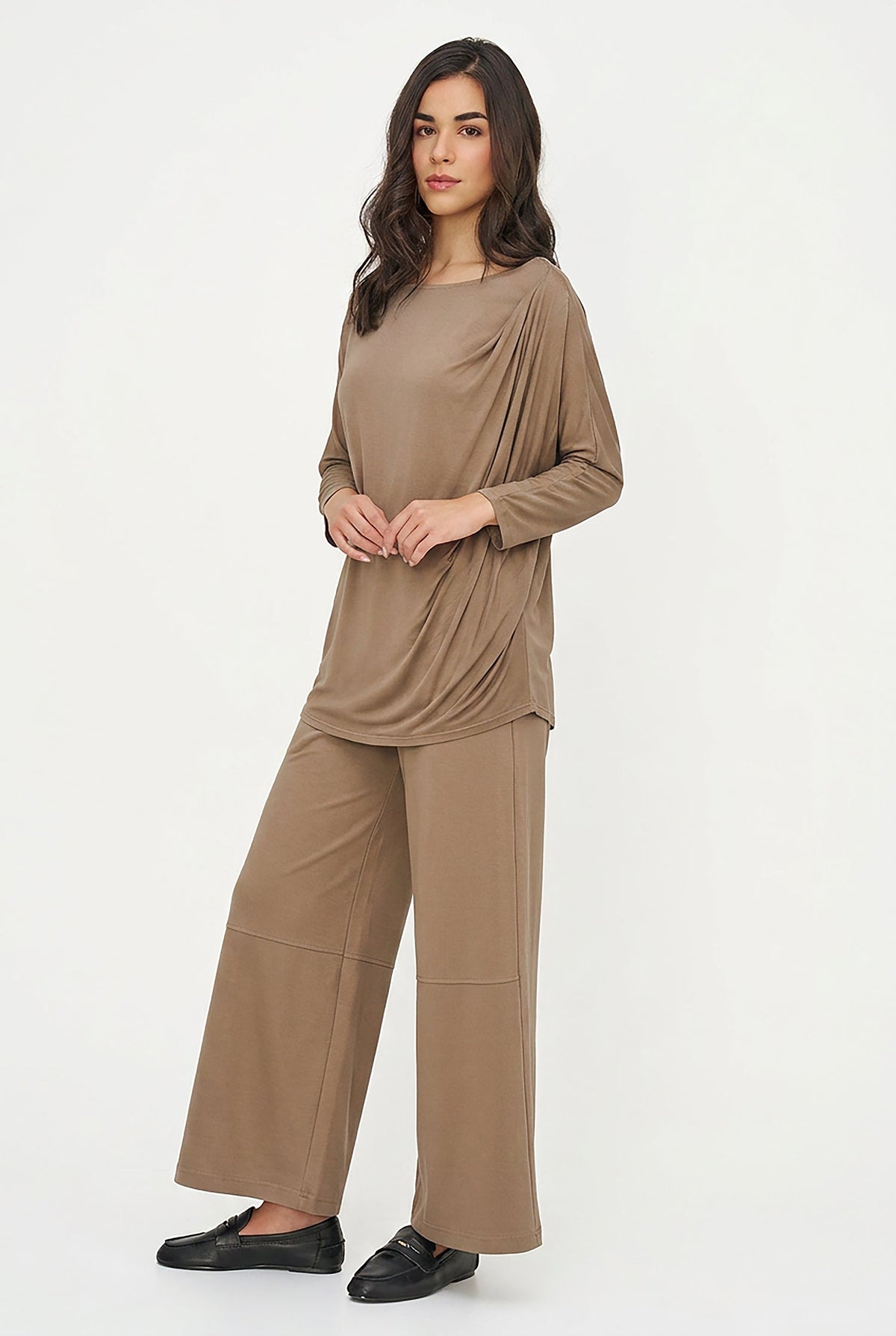 A woman modeling a matching two-piece lounge set in a warm taupe-brown color, featuring a draped long-sleeve top and wide-leg pants, paired with black loafers against a plain white studio background.