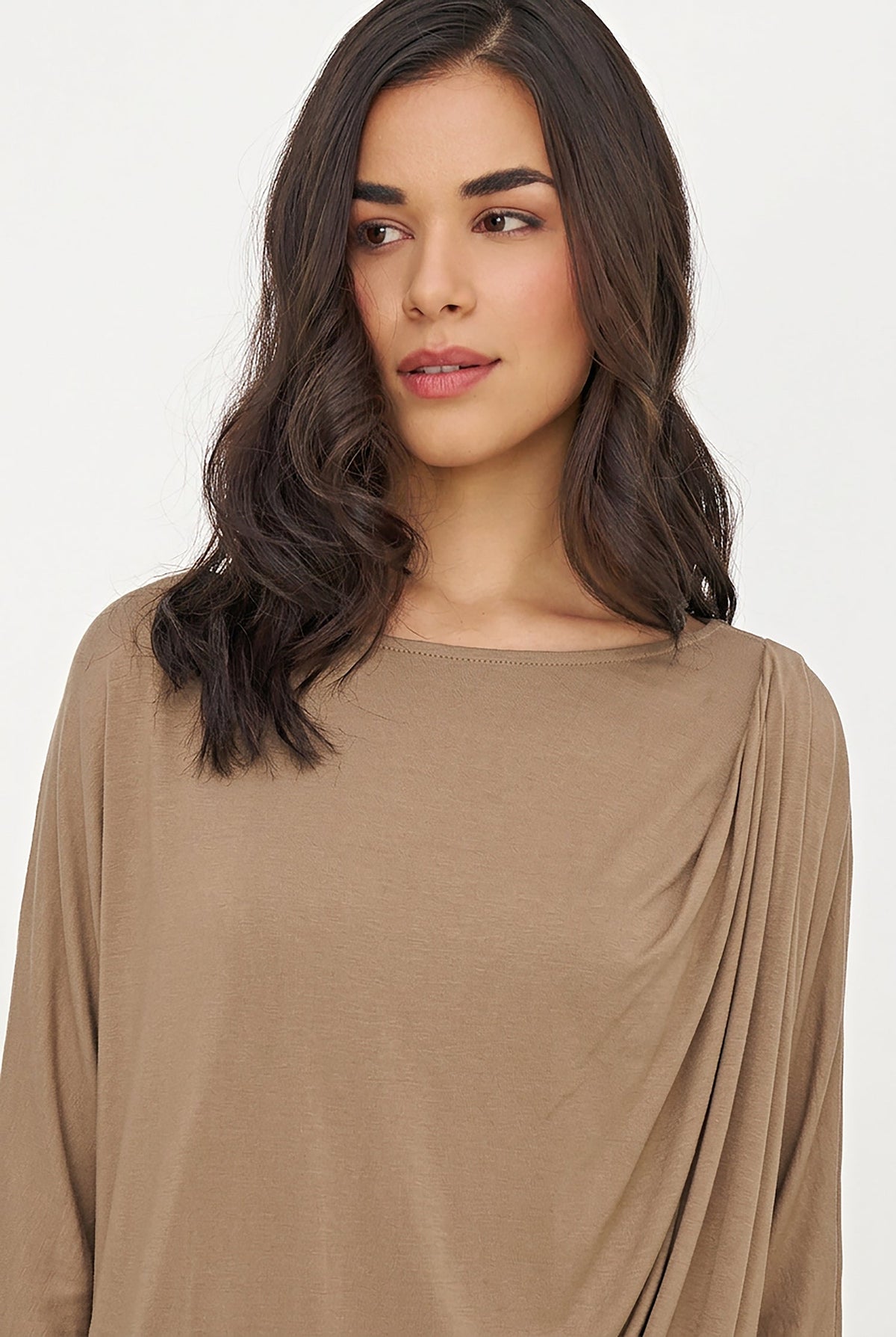 A woman with dark wavy hair wearing a draped, long-sleeve top in a warm taupe-brown color, photographed against a clean white background.