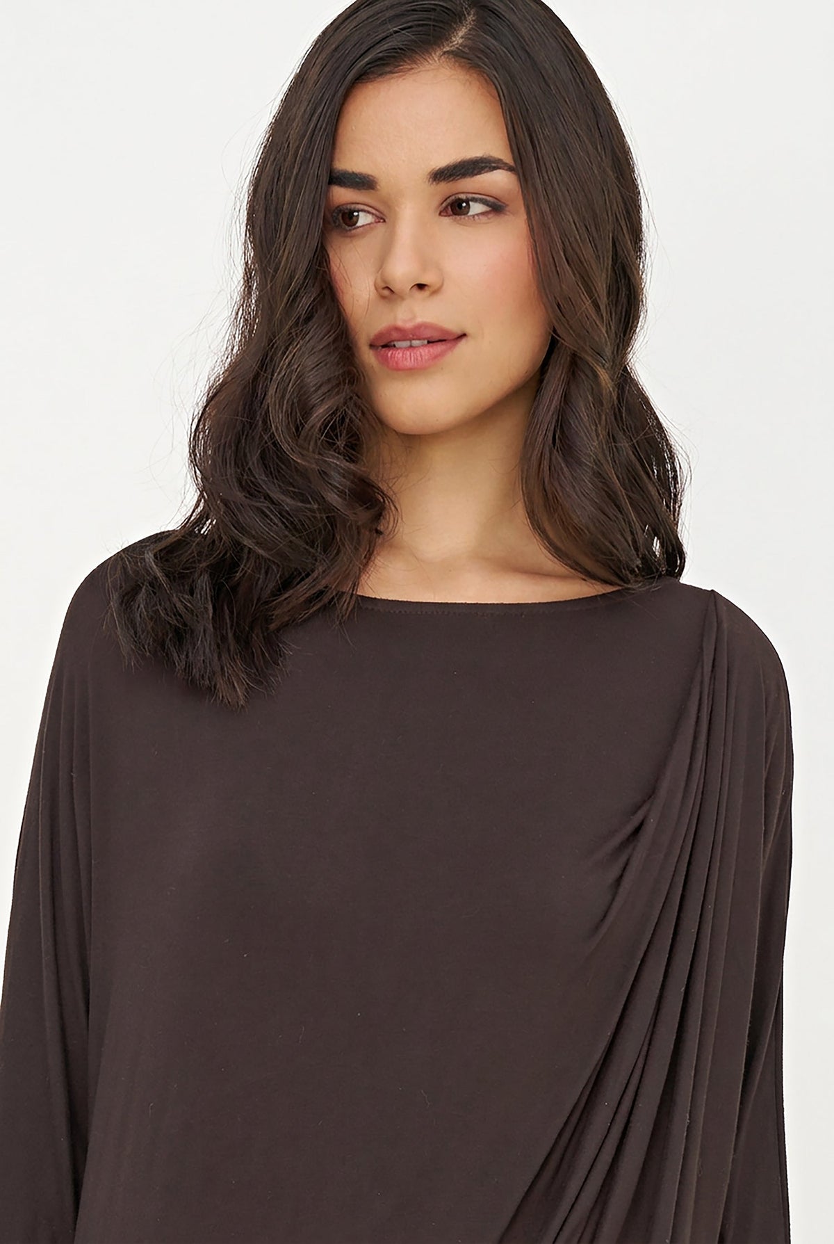 A woman with long dark brown hair wearing a dark brown draped long-sleeve top, shown from the chest up against a plain white background.