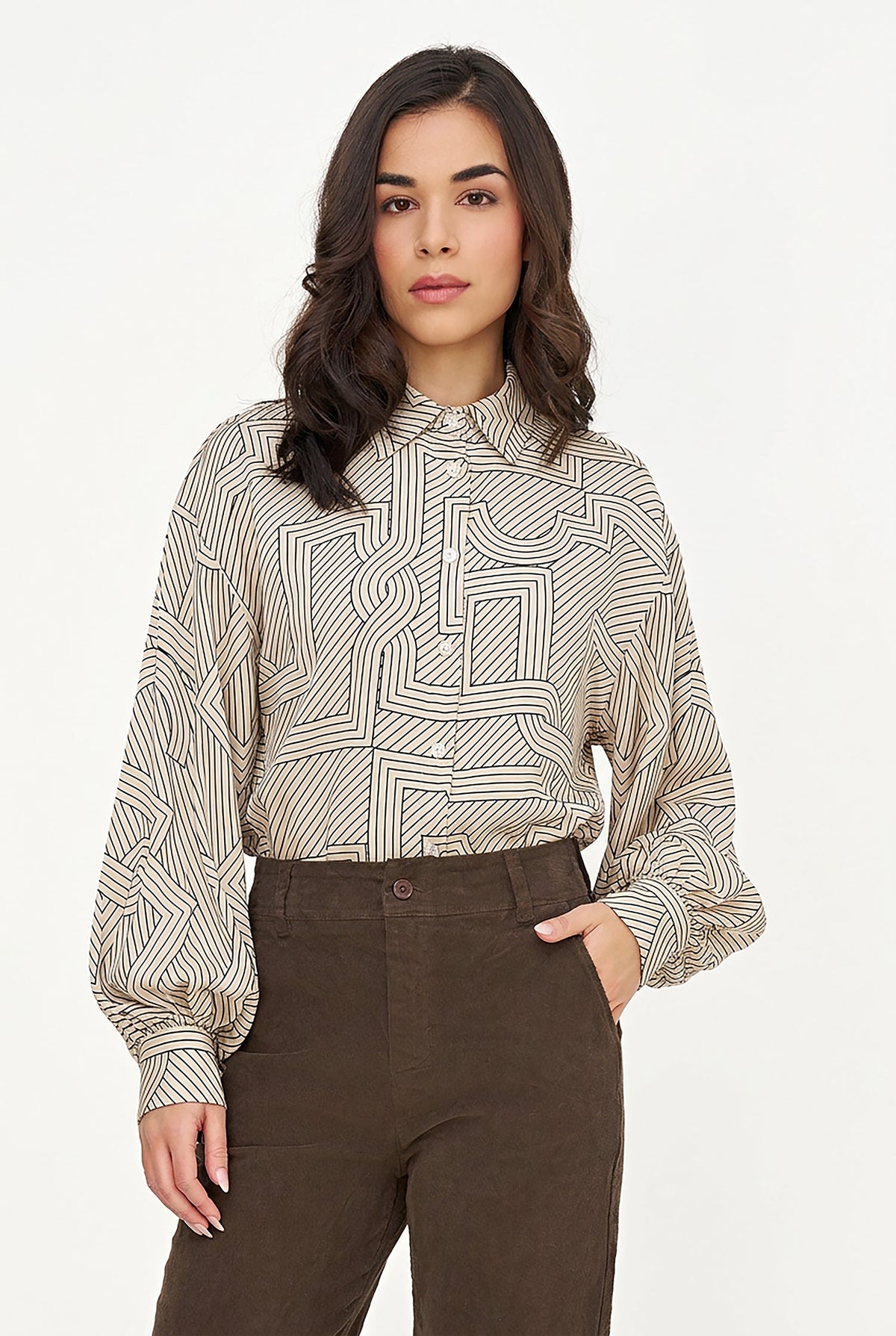 A woman modeling a beige and black patterned long-sleeve button-up shirt with black cropped trousers and brown suede loafers, standing against a plain white studio background.