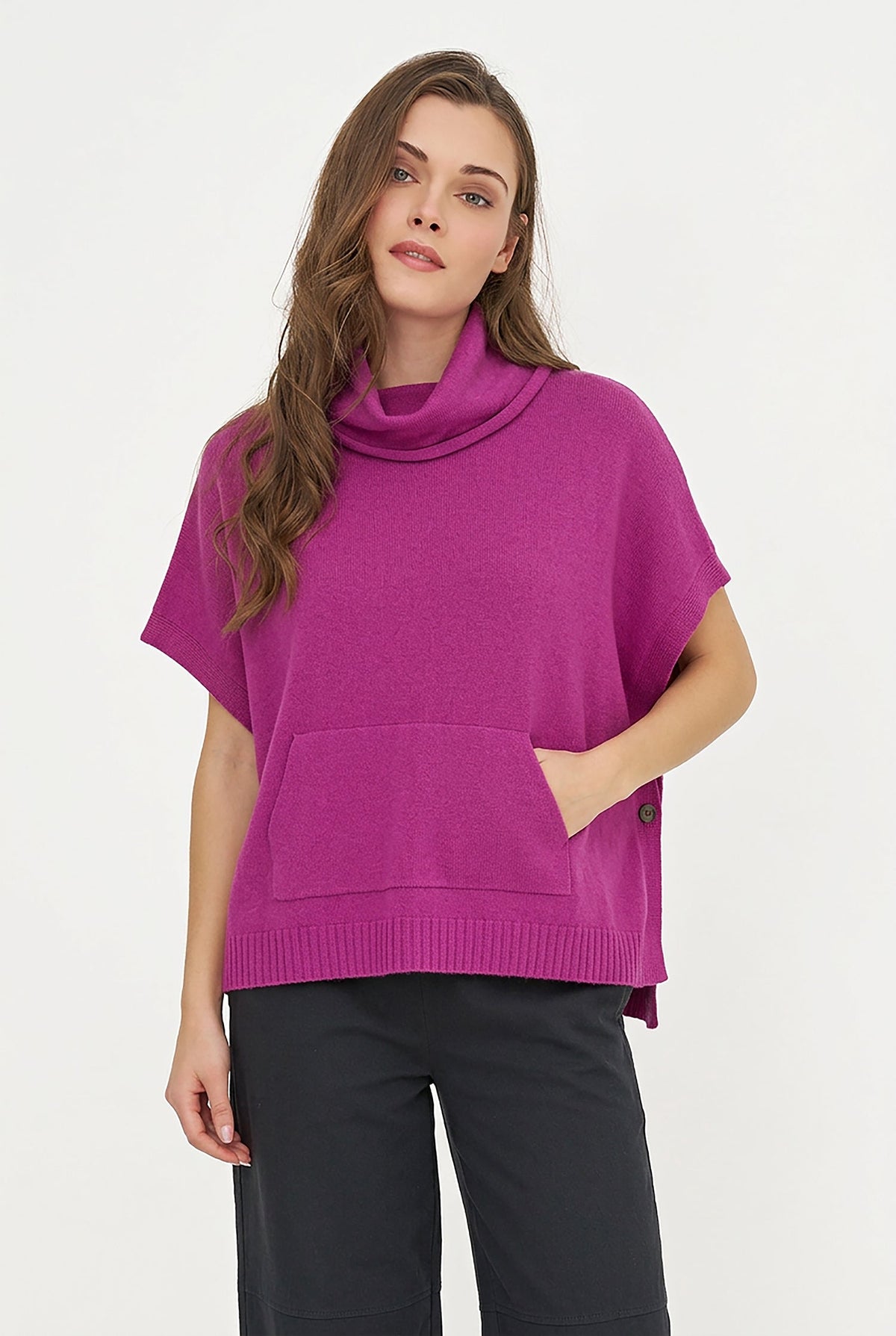 A woman wearing a bright magenta knit poncho sweater with a cowl neck and front pocket, paired with black pants, against a plain white studio background.