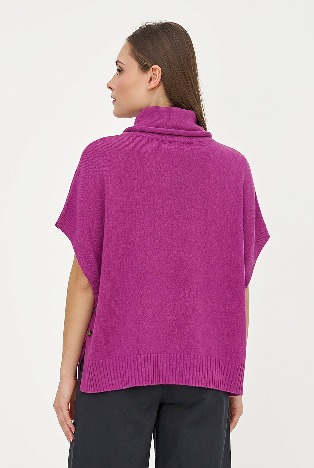 A woman modeling a bright magenta-purple knit cowl-neck poncho sweater with short sleeves and a front kangaroo pocket, paired with black pants, against a plain white studio background.