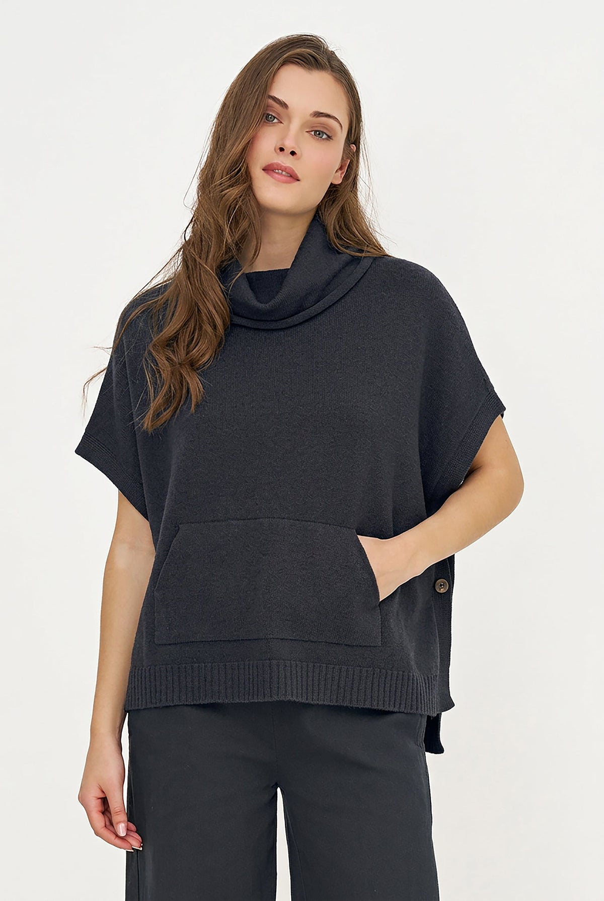 A woman modeling a black short-sleeve cowl-neck knit poncho sweater with a front patch pocket and ribbed hem, paired with black pants against a plain white studio background.