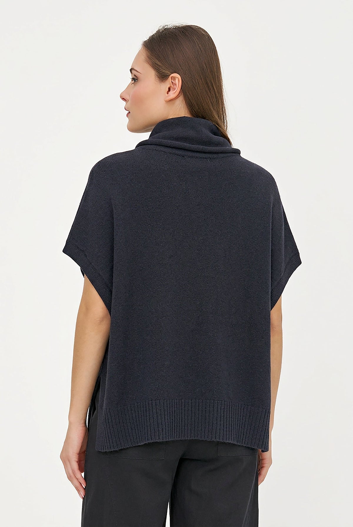 A woman modeling a dark charcoal gray cowl-neck poncho sweater with a front kangaroo pocket and ribbed hem, paired with dark trousers, against a plain light background.