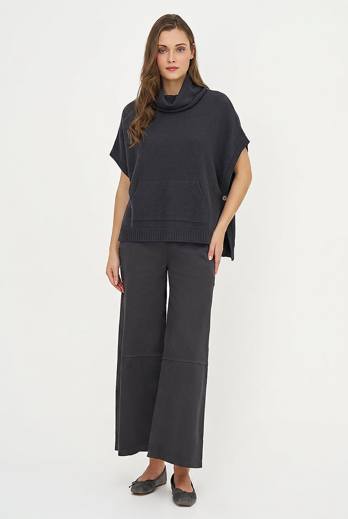 A woman modeling a dark charcoal gray knit set consisting of a short-sleeve cowl-neck poncho top and matching wide-leg cropped pants, paired with dark flat shoes, against a plain white studio background.