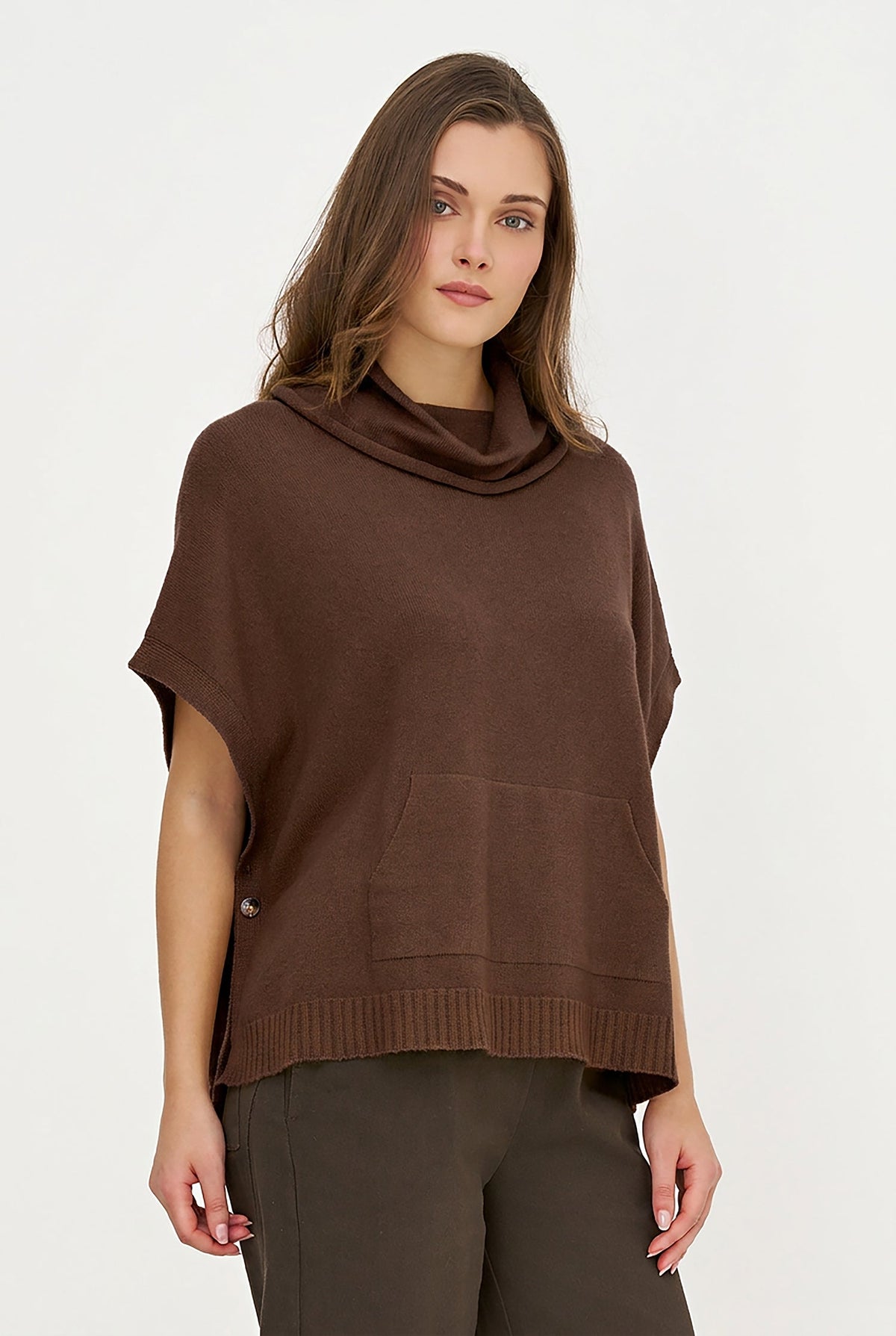 A woman wearing a brown cowl-neck knit poncho top with short sleeves and a front kangaroo pocket, paired with dark olive pants, against a plain white studio background.