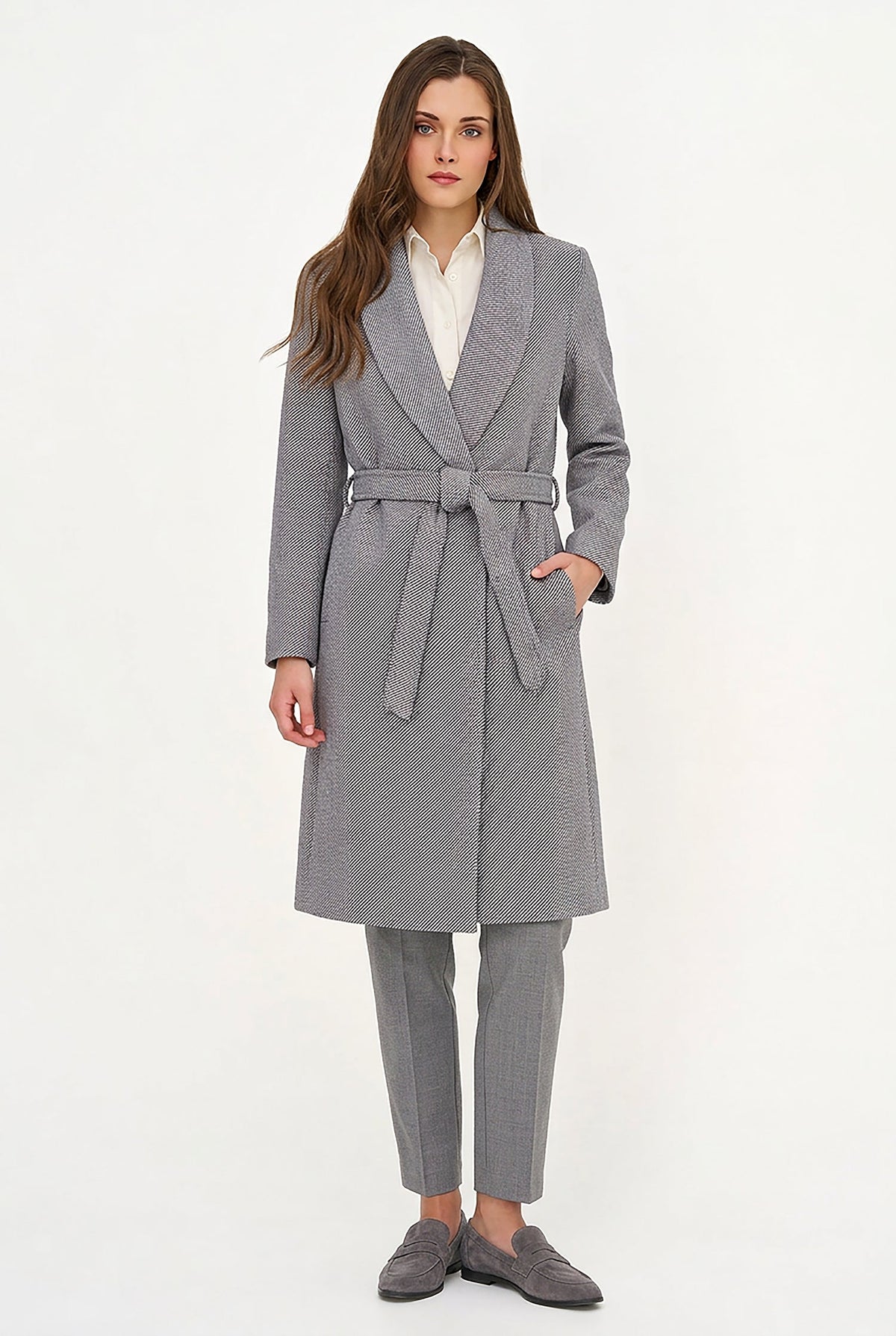 A woman modeling a gray belted coat over a white collared shirt, gray trousers, and gray loafers, photographed against a plain white studio background.