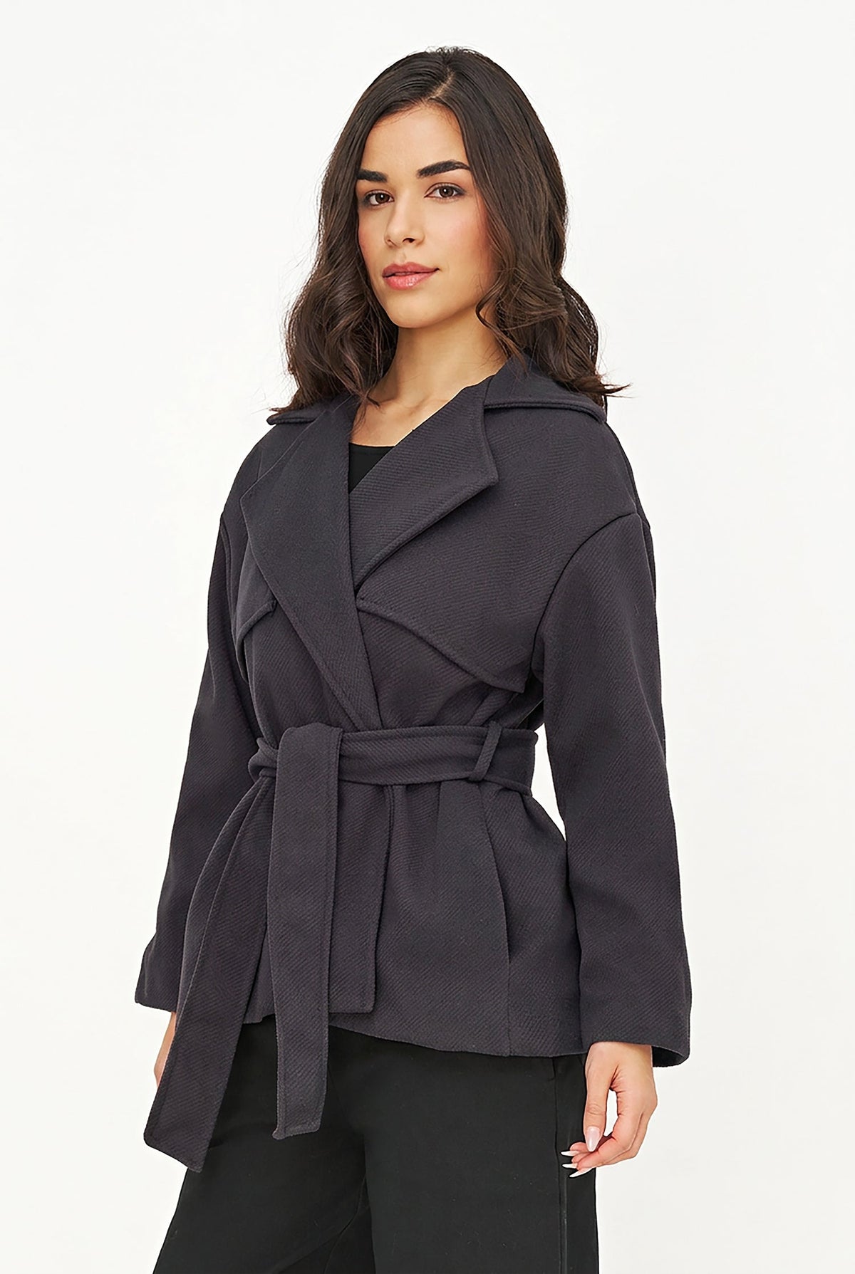 A woman modeling a dark charcoal gray belted wrap coat over a black top and black pants, photographed against a clean white studio background.