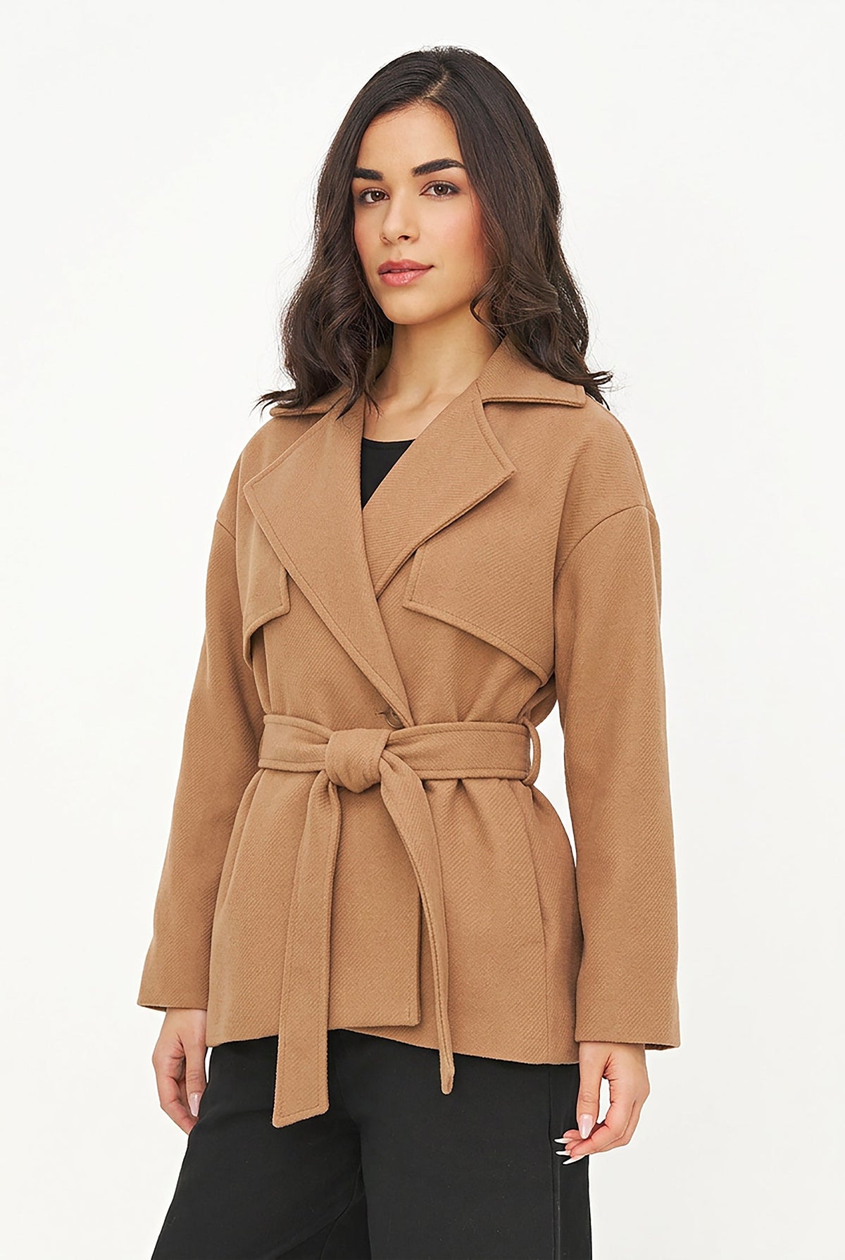 A woman modeling a camel brown belted wrap coat over a black top and black pants, photographed against a clean white studio background.