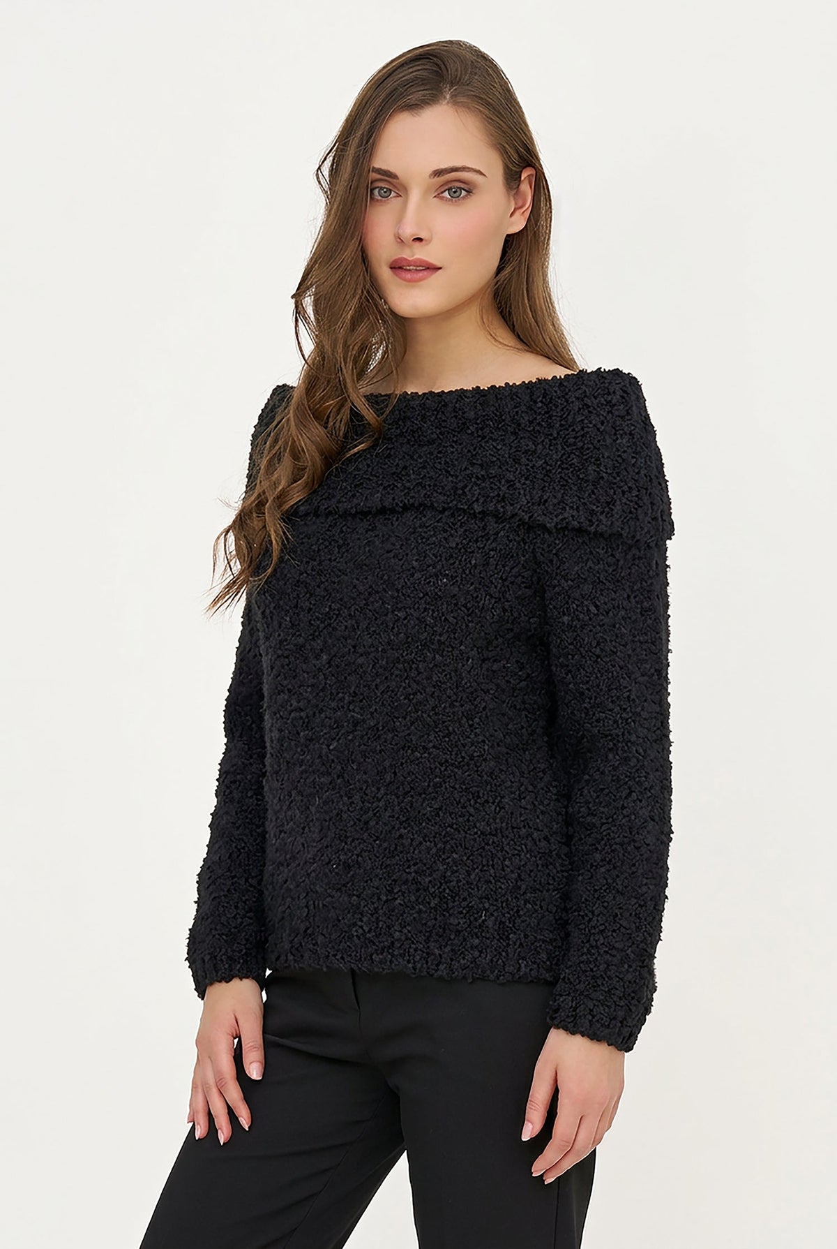 A woman modeling a black textured off-the-shoulder sweater with long sleeves, paired with black pants, against a plain white studio background.