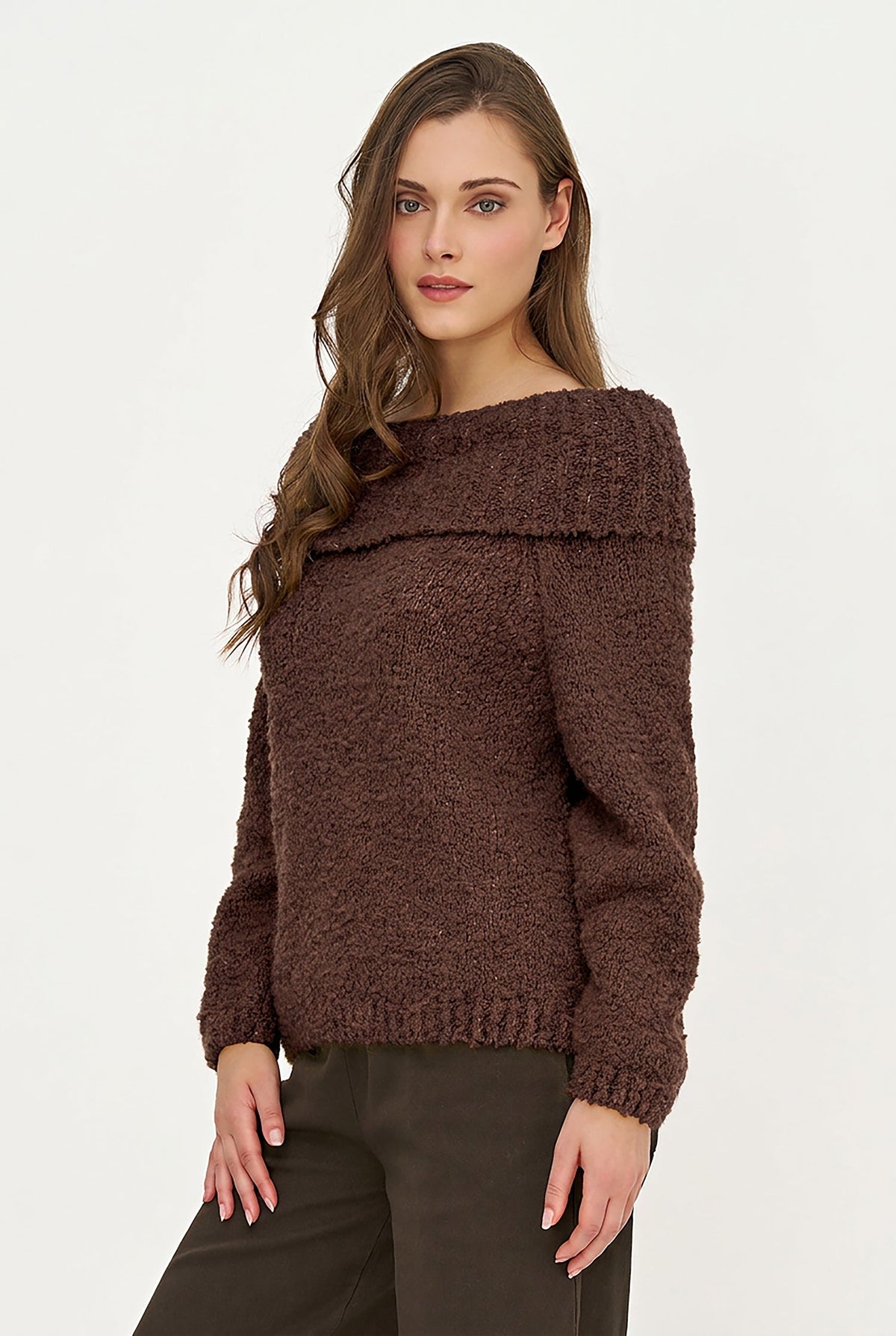 A woman modeling a brown textured off-the-shoulder knit sweater with long sleeves, paired with dark brown pants, against a plain white studio background.