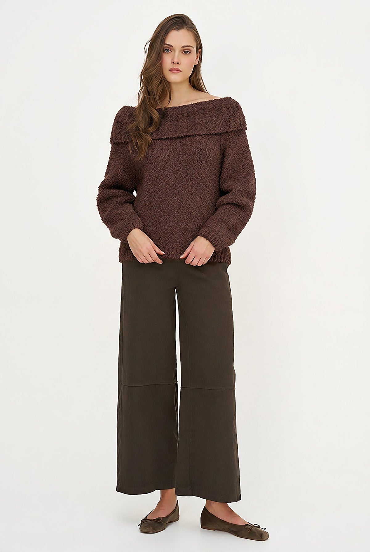A woman modeling a dark brown textured off-the-shoulder sweater paired with dark brown wide-leg trousers and brown flats, set against a plain white studio background.