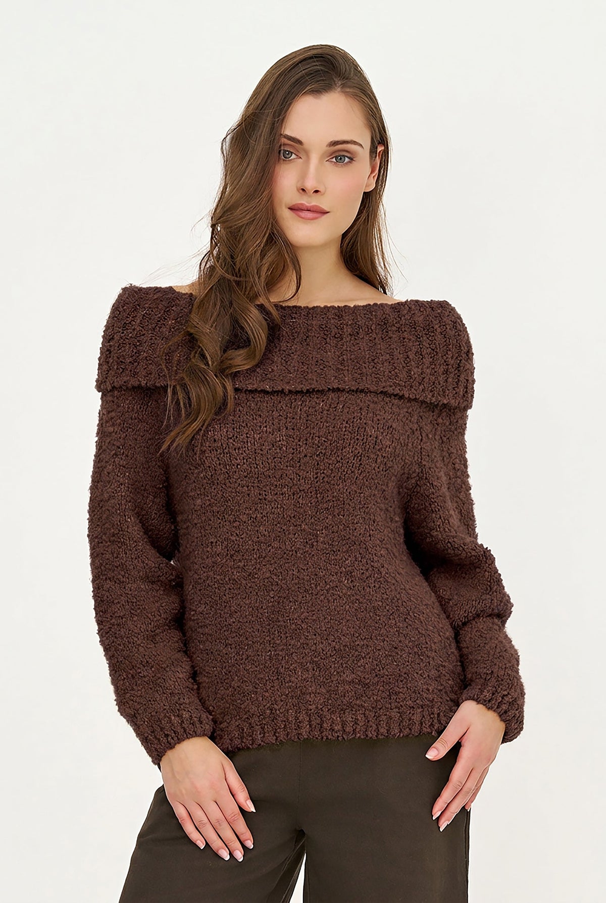 A woman modeling a chunky knit off-the-shoulder sweater in a dark brown color, paired with dark brown pants, against a plain white studio background.