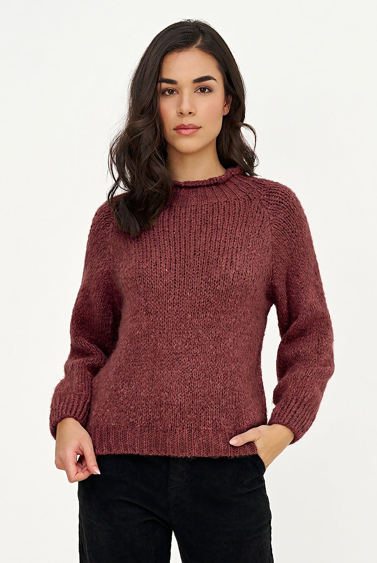 A woman modeling a chunky, ribbed knit crewneck sweater in a dusty rose or mauve color, paired with black pants, against a plain white studio background.