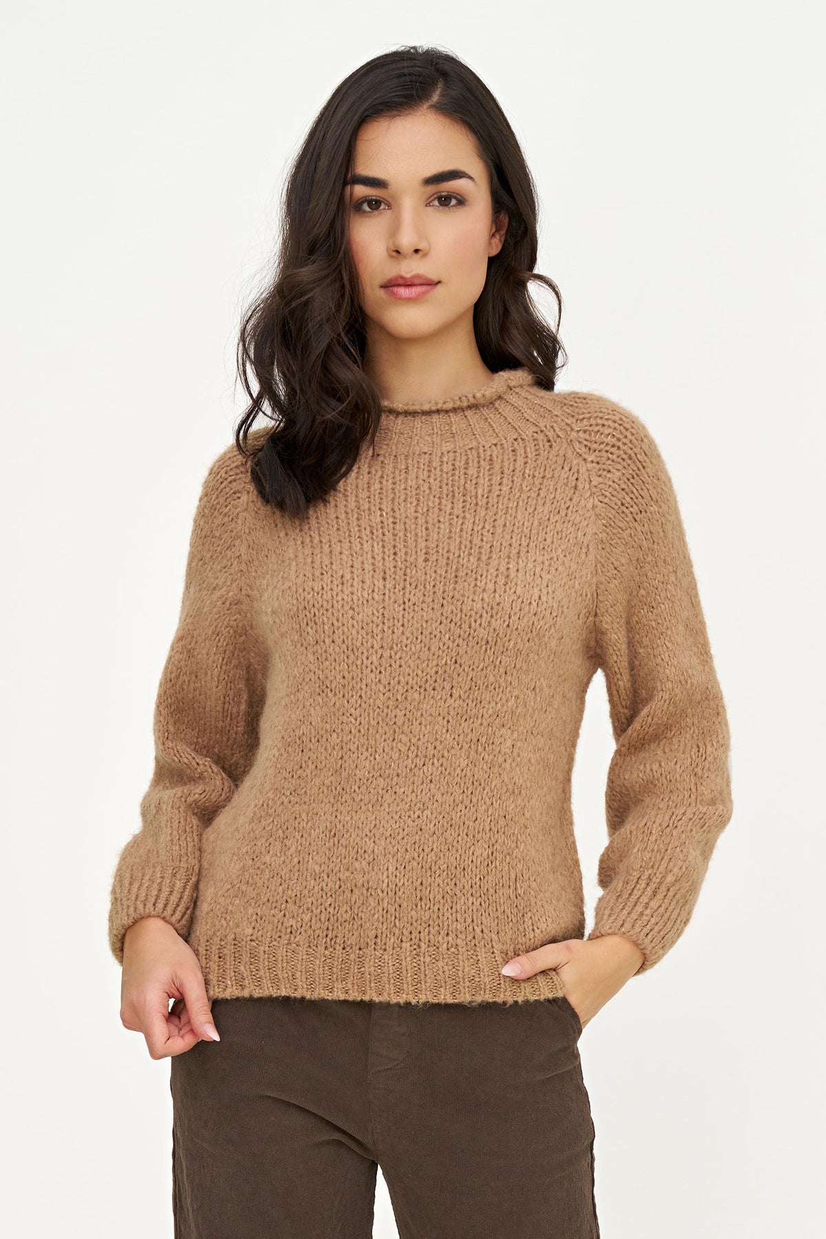 A woman modeling a cozy, ribbed knit crewneck sweater in a warm camel brown color, paired with dark brown pants, against a plain white studio background.