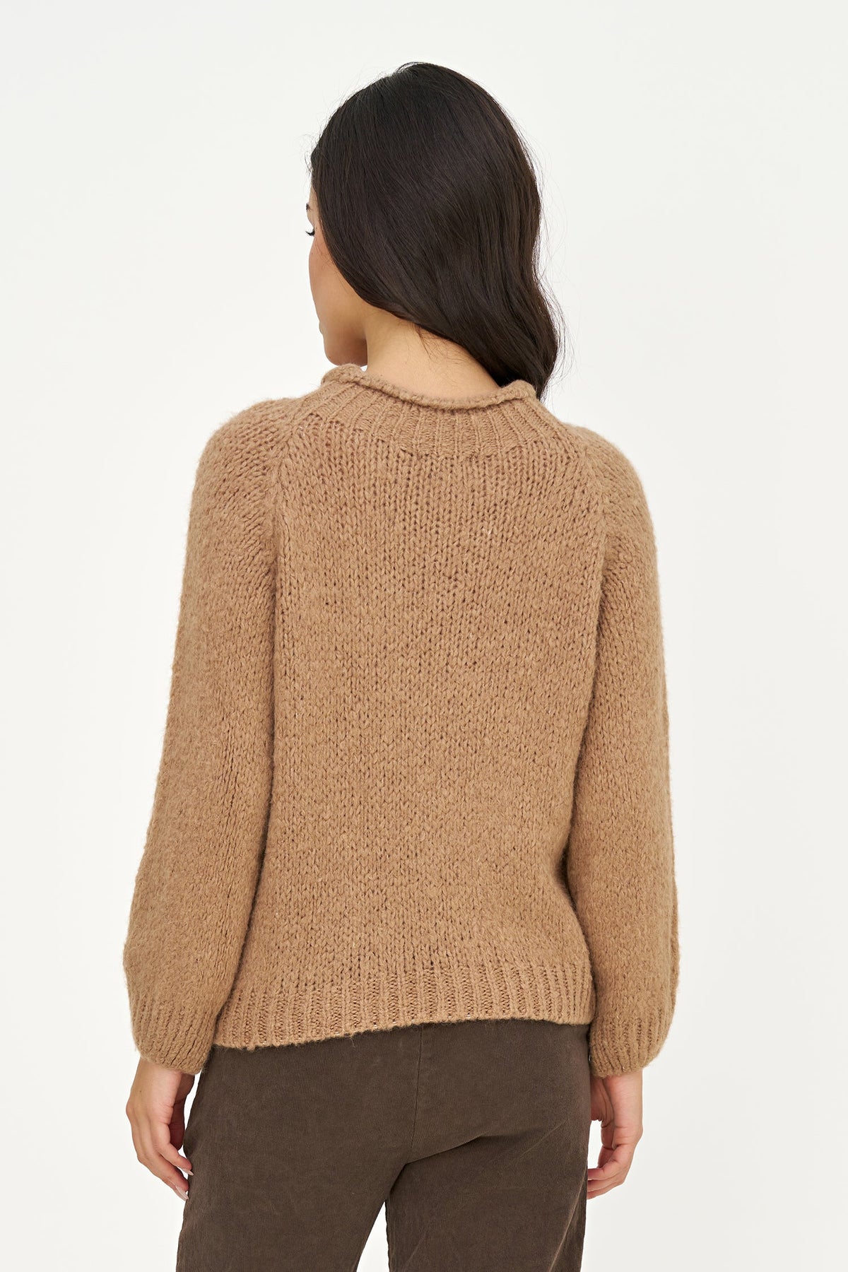A woman viewed from the back wearing a textured, chunky knit sweater in a warm camel brown color, paired with dark brown pants, against a plain white studio background.