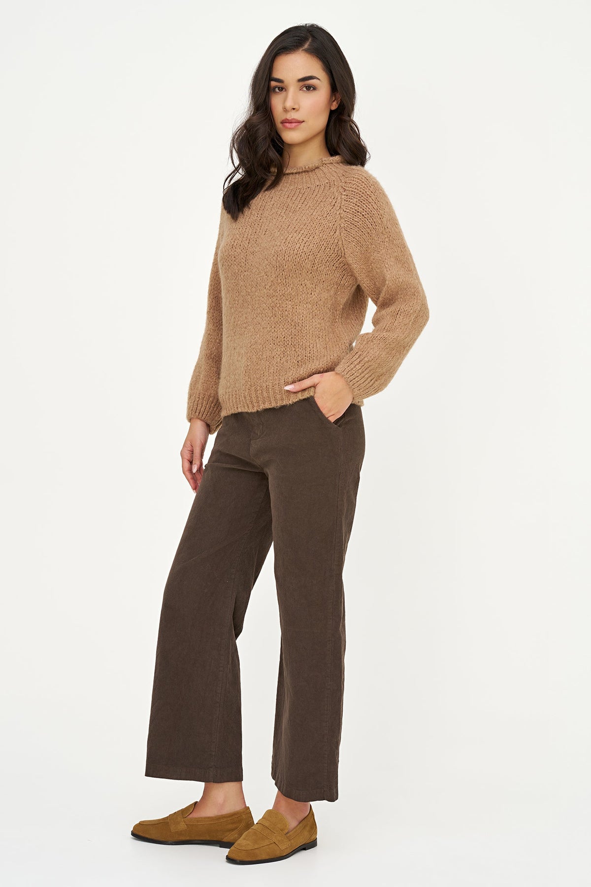 A woman modeling a beige chunky knit crewneck sweater and dark brown wide-leg trousers, paired with brown suede loafers, against a plain white studio background.