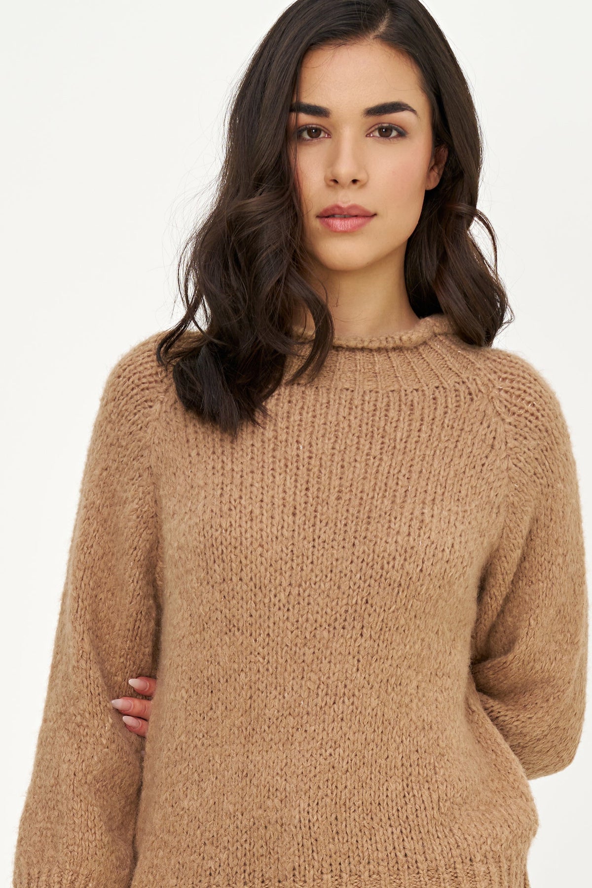 A woman wearing a chunky, ribbed knit crewneck sweater in a warm camel brown color, photographed against a clean white studio background.