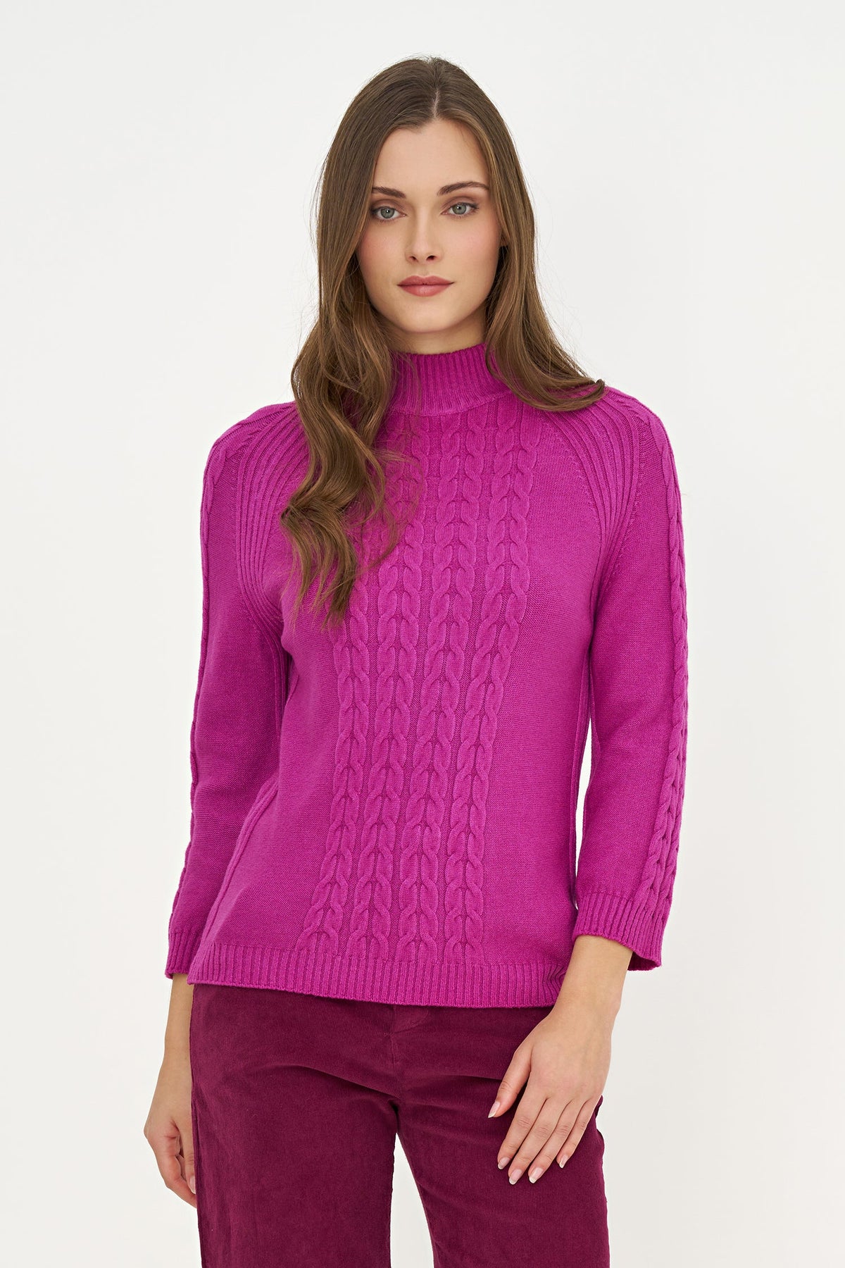 A woman modeling a bright magenta cable-knit mock neck sweater with ribbed cuffs and hem, paired with matching corduroy trousers, against a plain white studio background.
