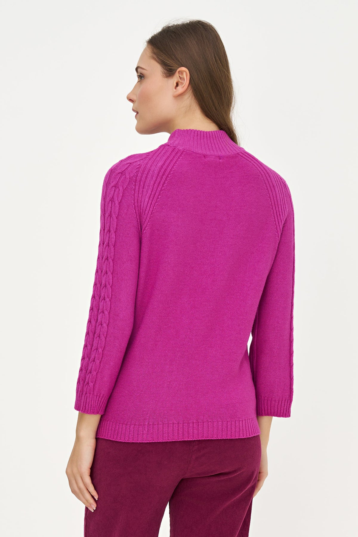 A woman shown from the back wearing a bright magenta knit sweater with cable detailing on the sleeves and matching dark magenta trousers, against a plain white studio background.