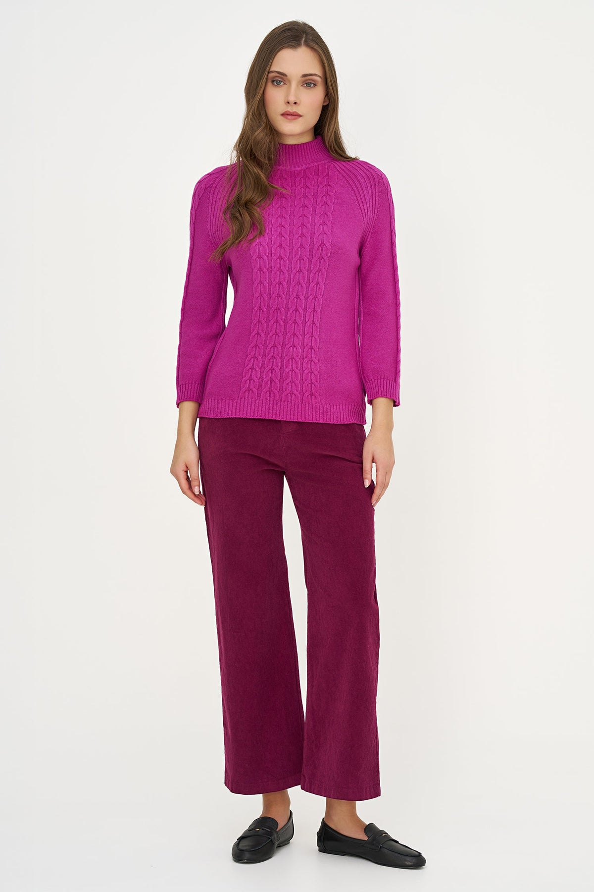 A woman modeling a bright magenta cable-knit mock-neck sweater paired with deep magenta corduroy trousers and black loafers, shot against a plain white studio background.