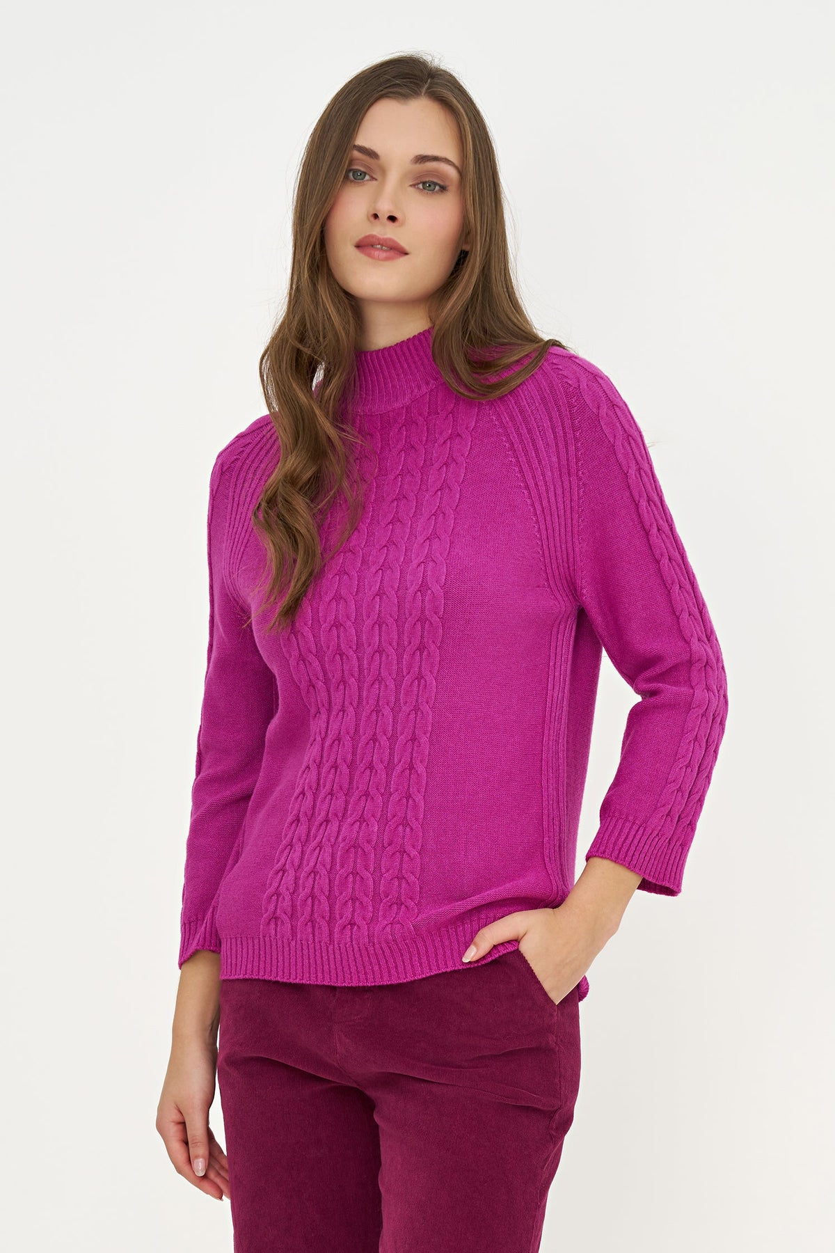 A woman wearing a bright magenta cable-knit mock-neck sweater and matching corduroy trousers, standing against a plain white studio background.