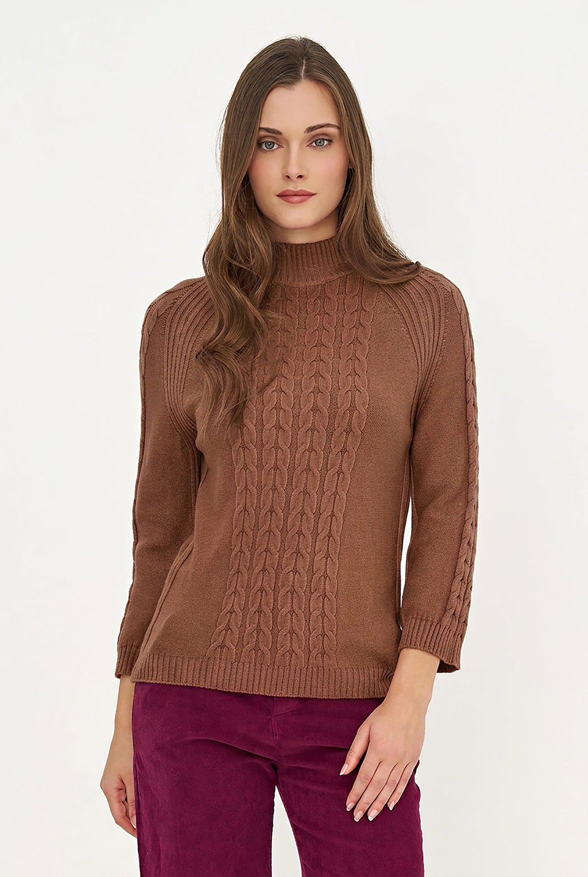 A woman modeling a brown cable-knit mock neck sweater with three-quarter sleeves, paired with magenta pants, against a plain white studio background.