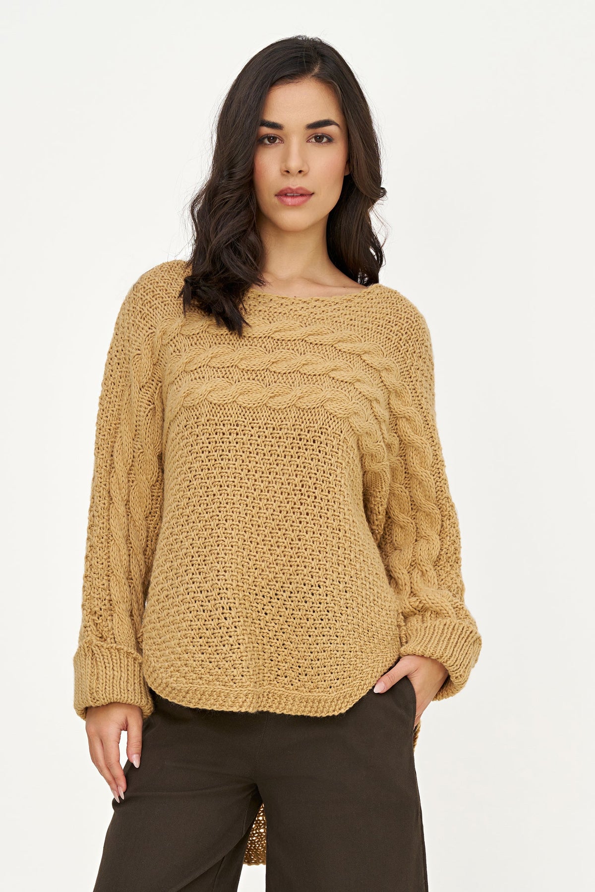 A woman modeling a chunky cable-knit sweater in a warm camel tan color, paired with dark brown pants, against a plain white studio background.