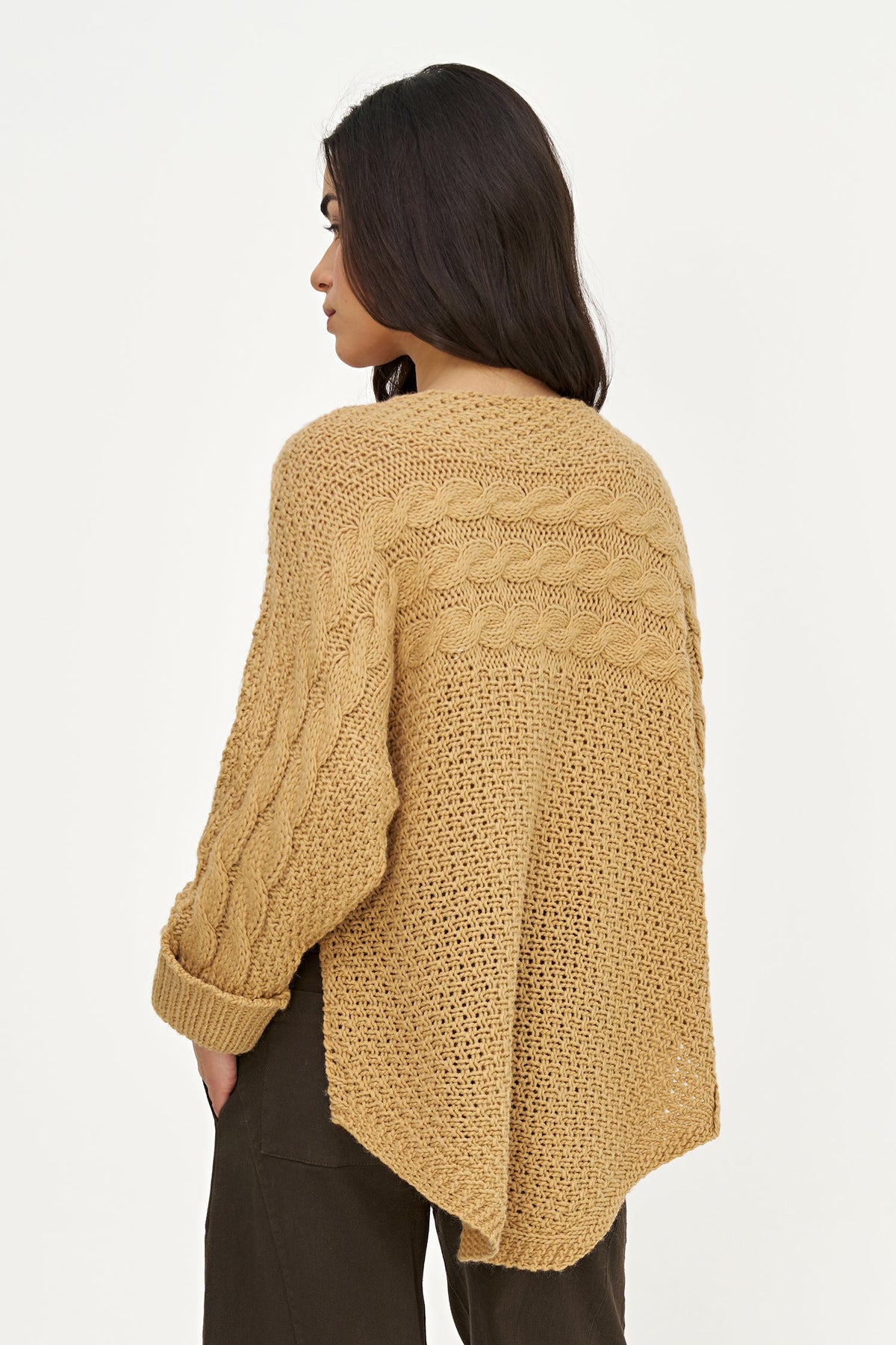 A woman shown from the back wearing a chunky, open-knit cable sweater in a warm camel tan color, paired with dark brown pants, against a plain white studio background.