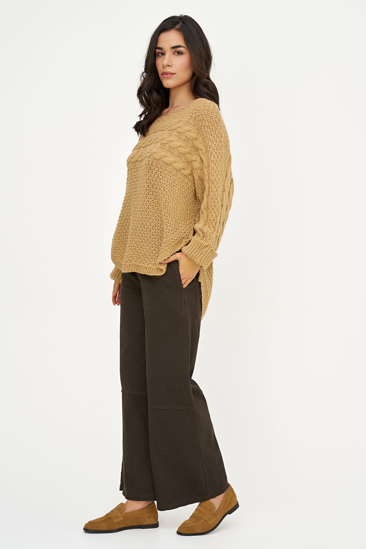 A woman modeling a chunky knit sweater in camel beige, paired with dark brown wide-leg trousers and tan suede loafers, against a plain white studio background.