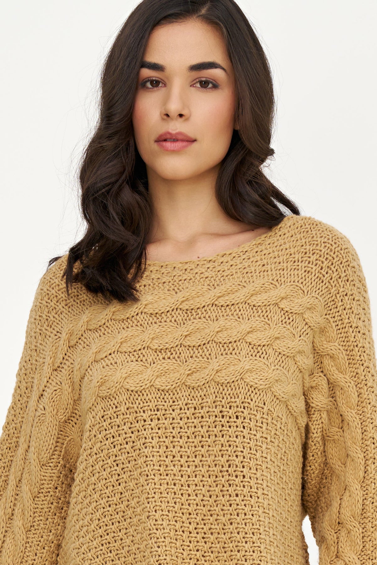 A woman wearing a chunky cable-knit sweater in a warm camel tan color, shown from the chest up against a plain light background.