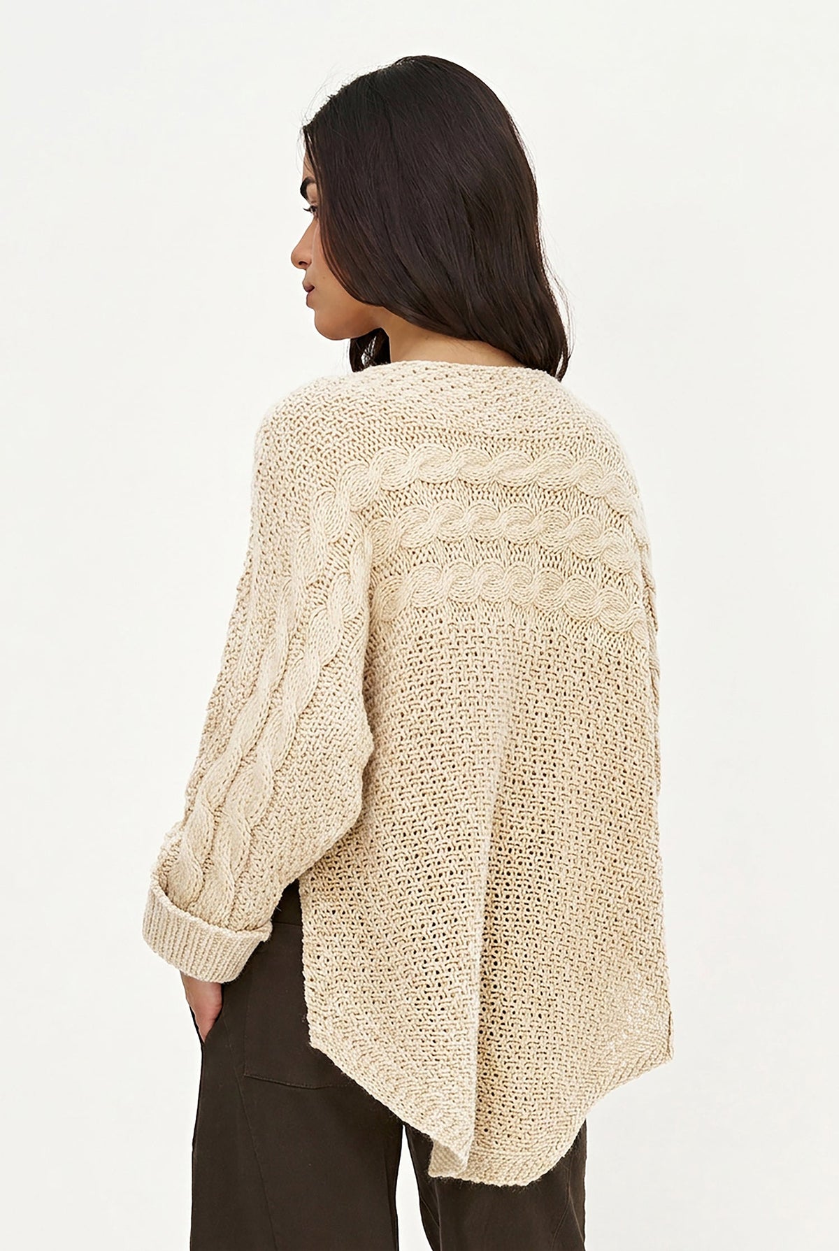 A woman shown from the back wearing a cream cable-knit cropped sweater and dark brown pants against a plain white studio background.