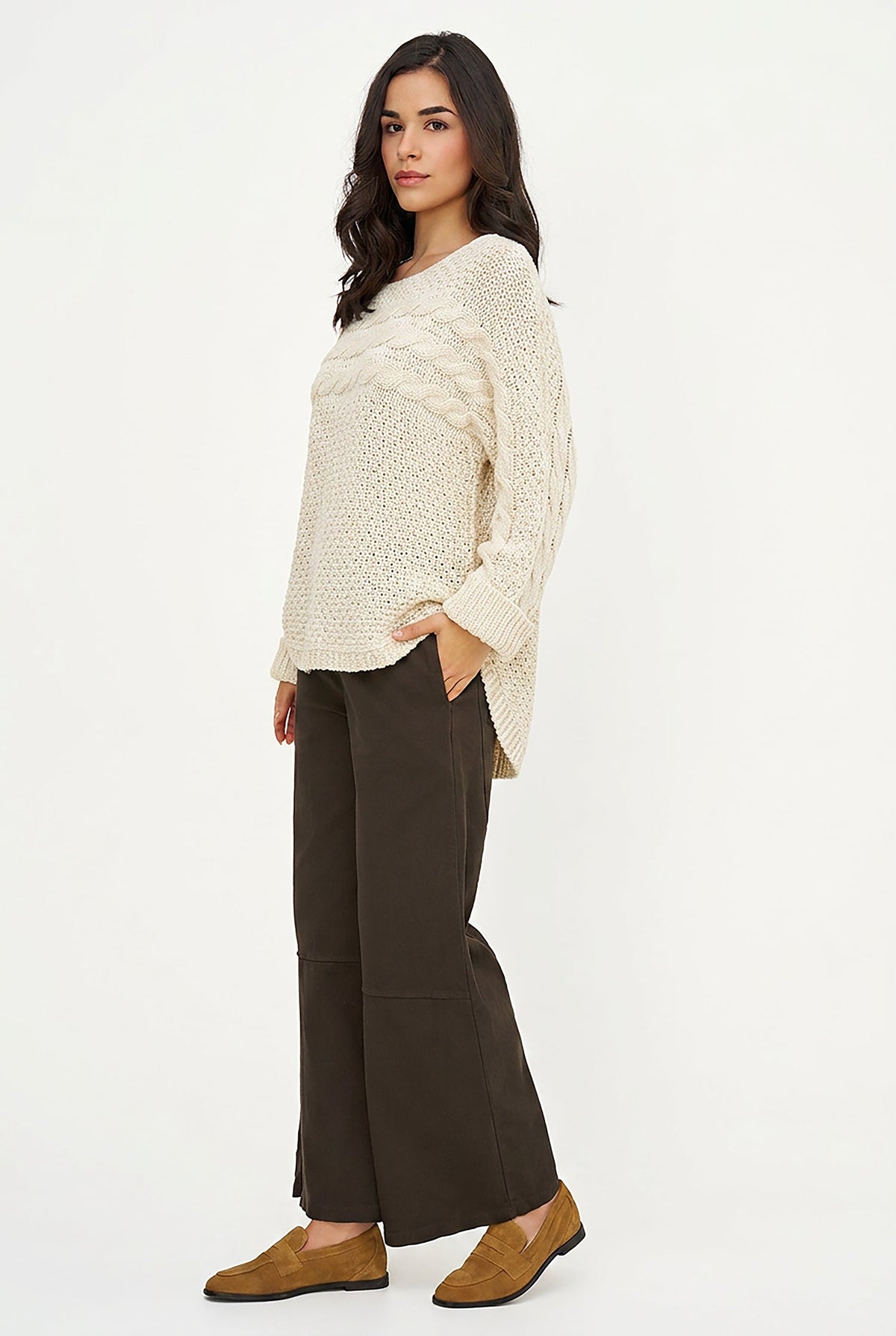 A woman modeling a cream cable-knit long-sleeve sweater, dark brown wide-leg cropped pants, and tan suede loafers against a plain white studio background.