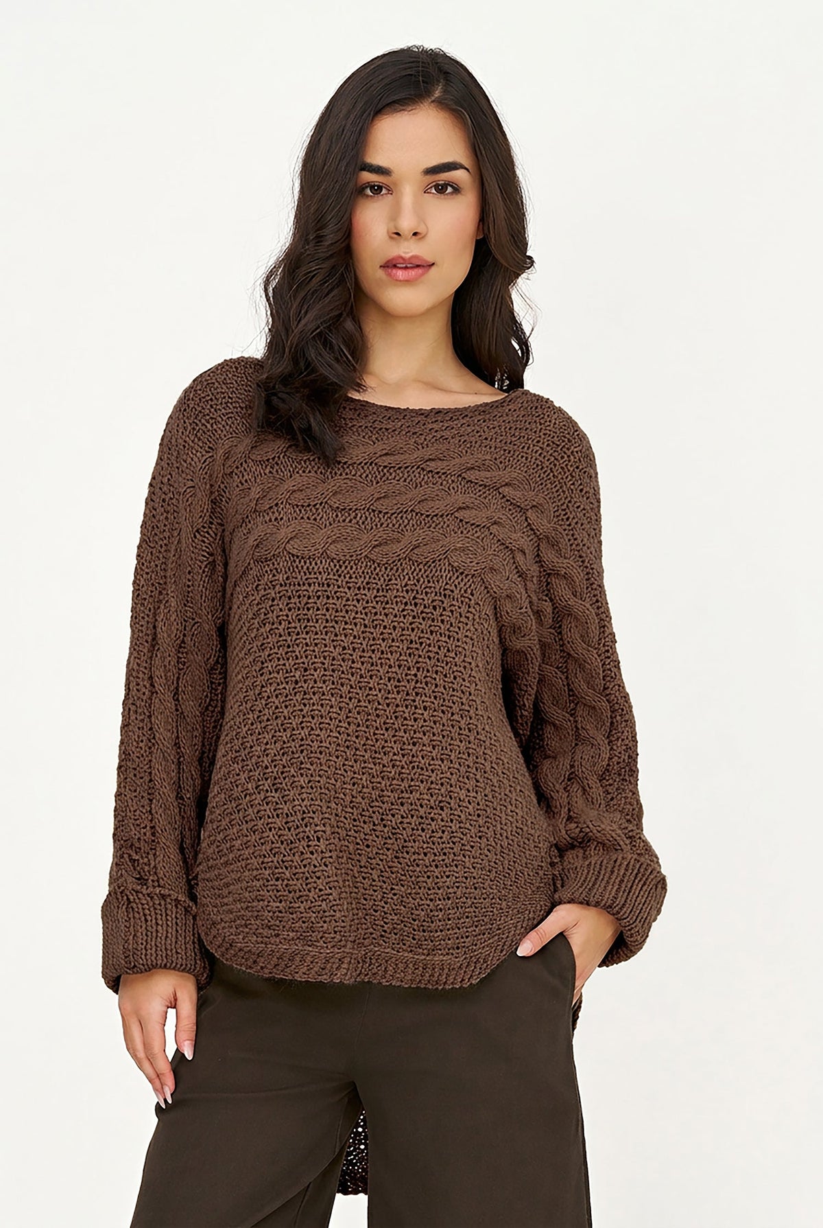 A woman modeling a chunky brown cable-knit sweater with a boat neckline, paired with dark brown pants, against a plain white studio background.