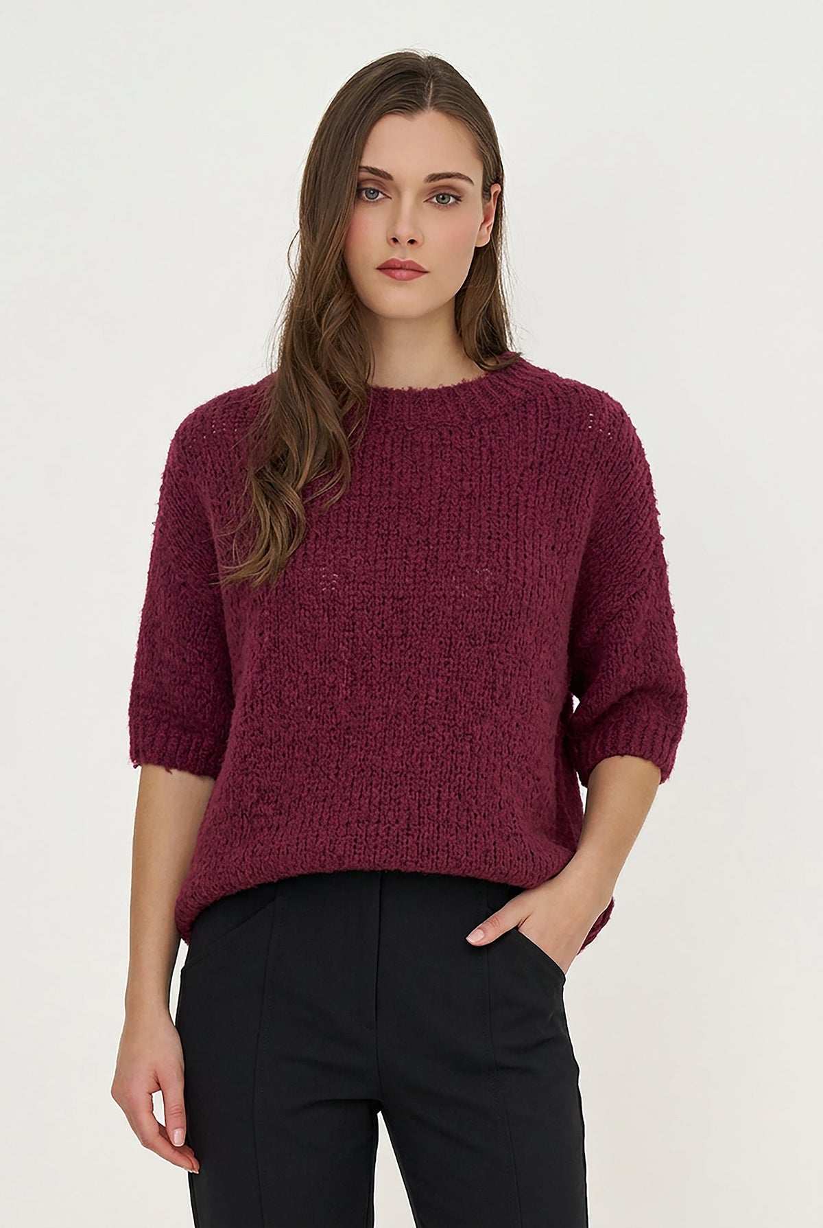 A woman modeling a textured, chunky knit short-sleeve sweater in a deep berry purple, paired with black trousers, against a plain white studio background.